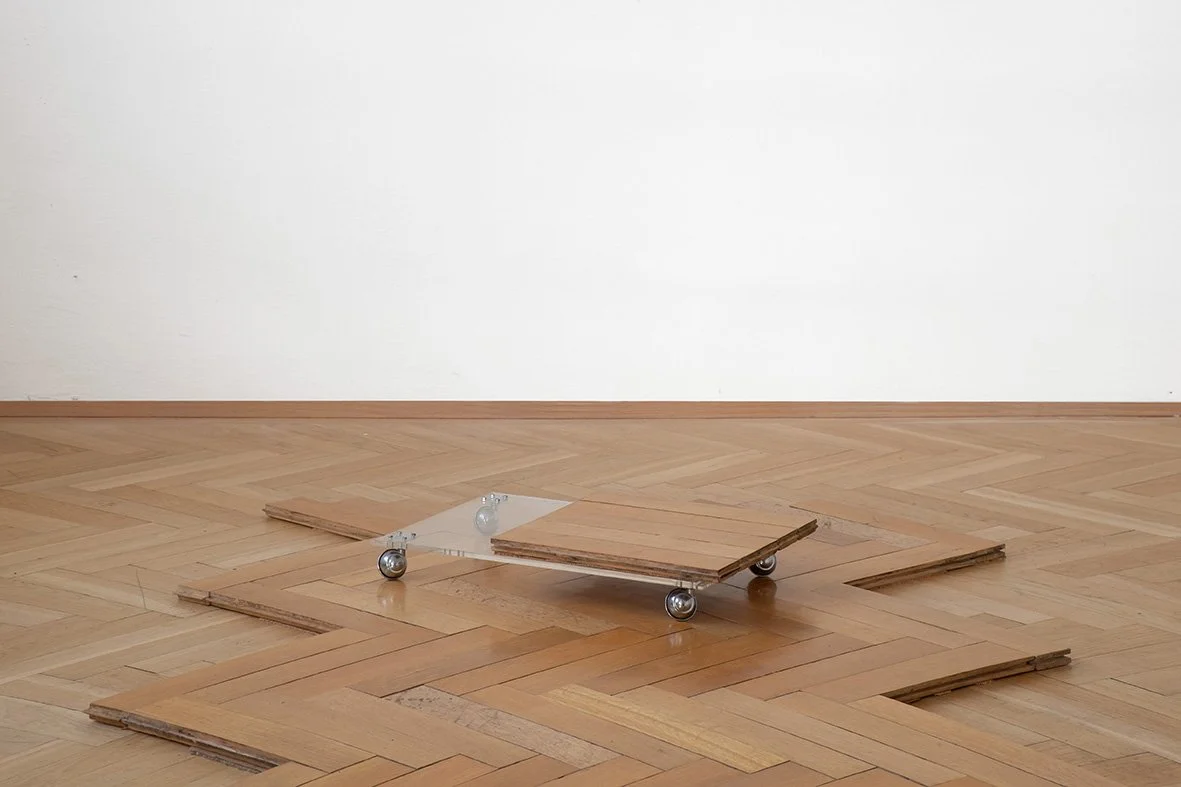 An art installation featuring a skateboard with a clear acrylic ramp on top, placed on a wooden parquet floor with several wooden planks arranged around it.