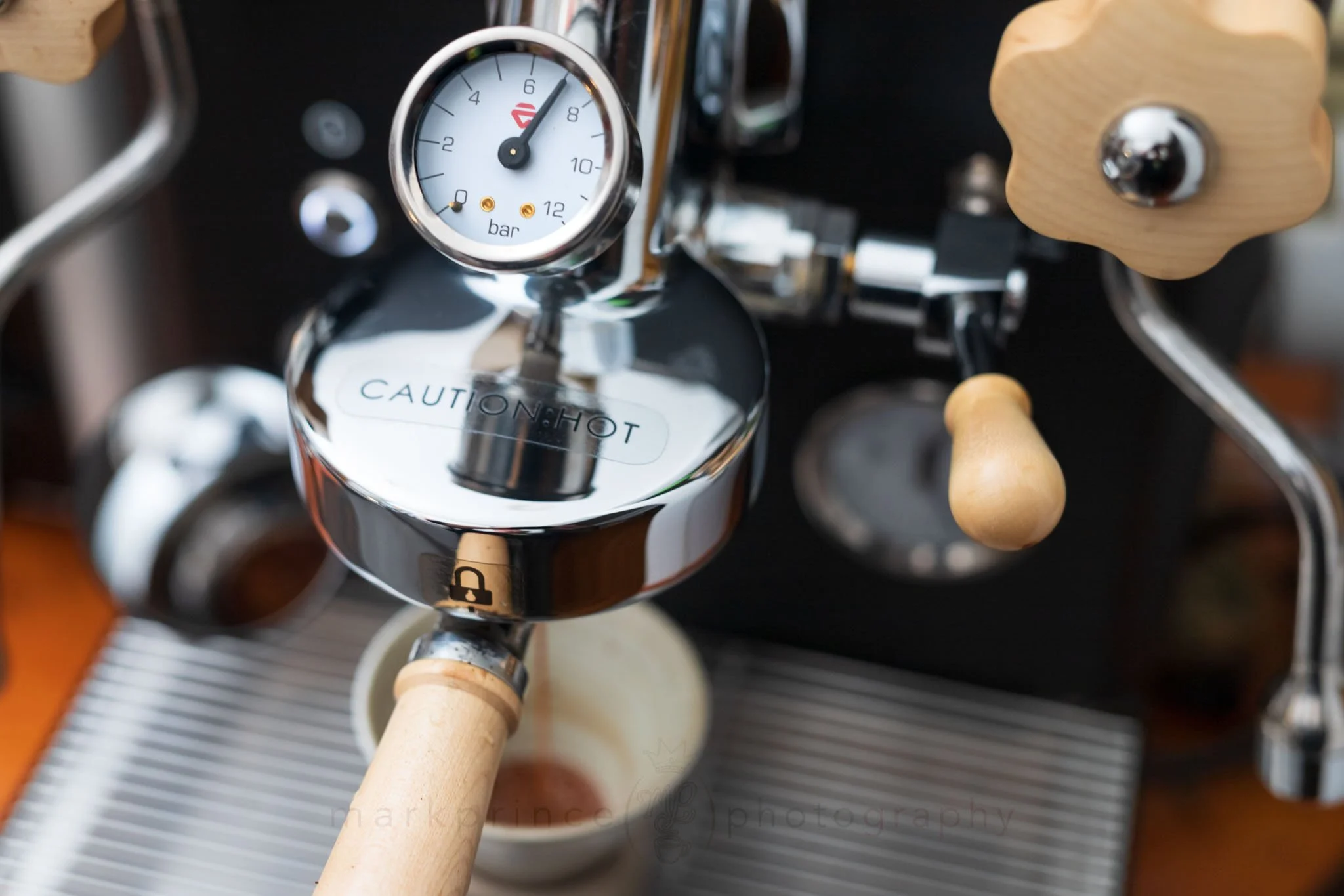 Close-up of a shiny espresso machine with a pressure gauge showing about 8 bar, wooden handles, and a warning label that reads 'CAUTION: HOT'.