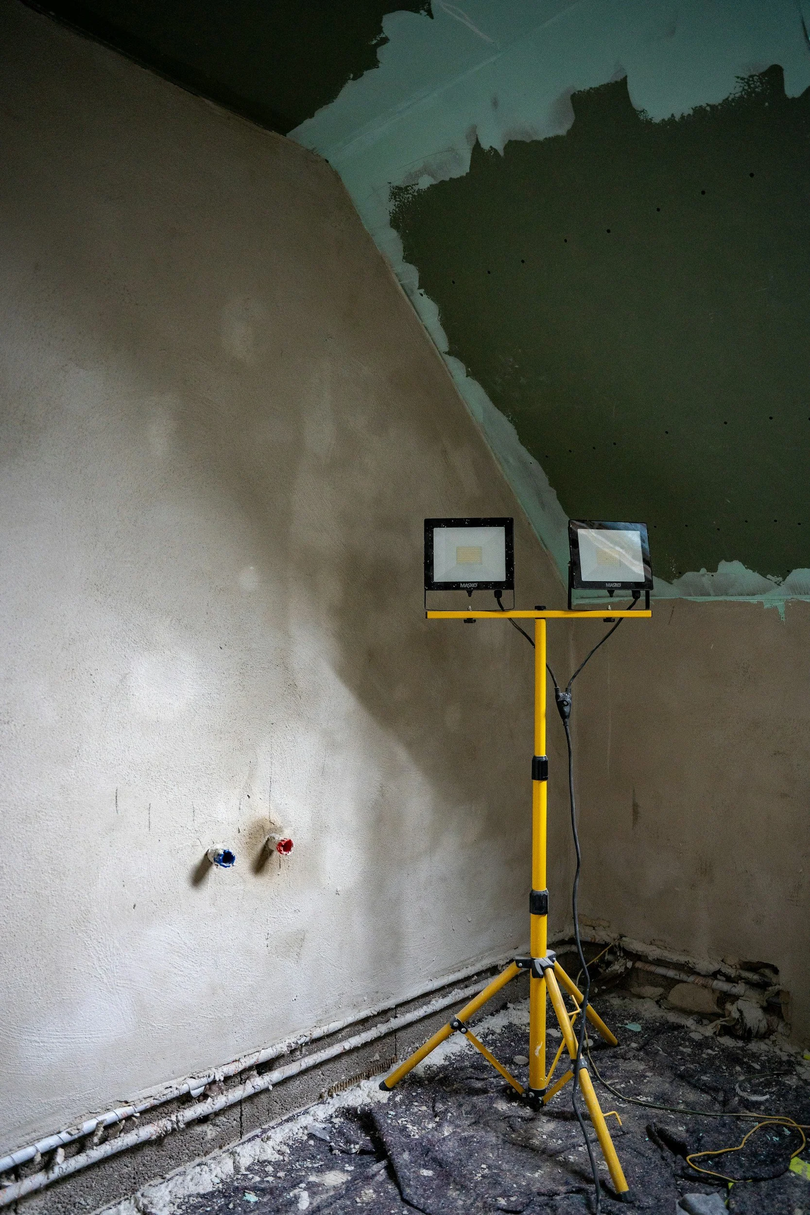 Construction site with unfinished walls and ceiling, two LED work lights on a yellow tripod, and exposed wiring and pipes.