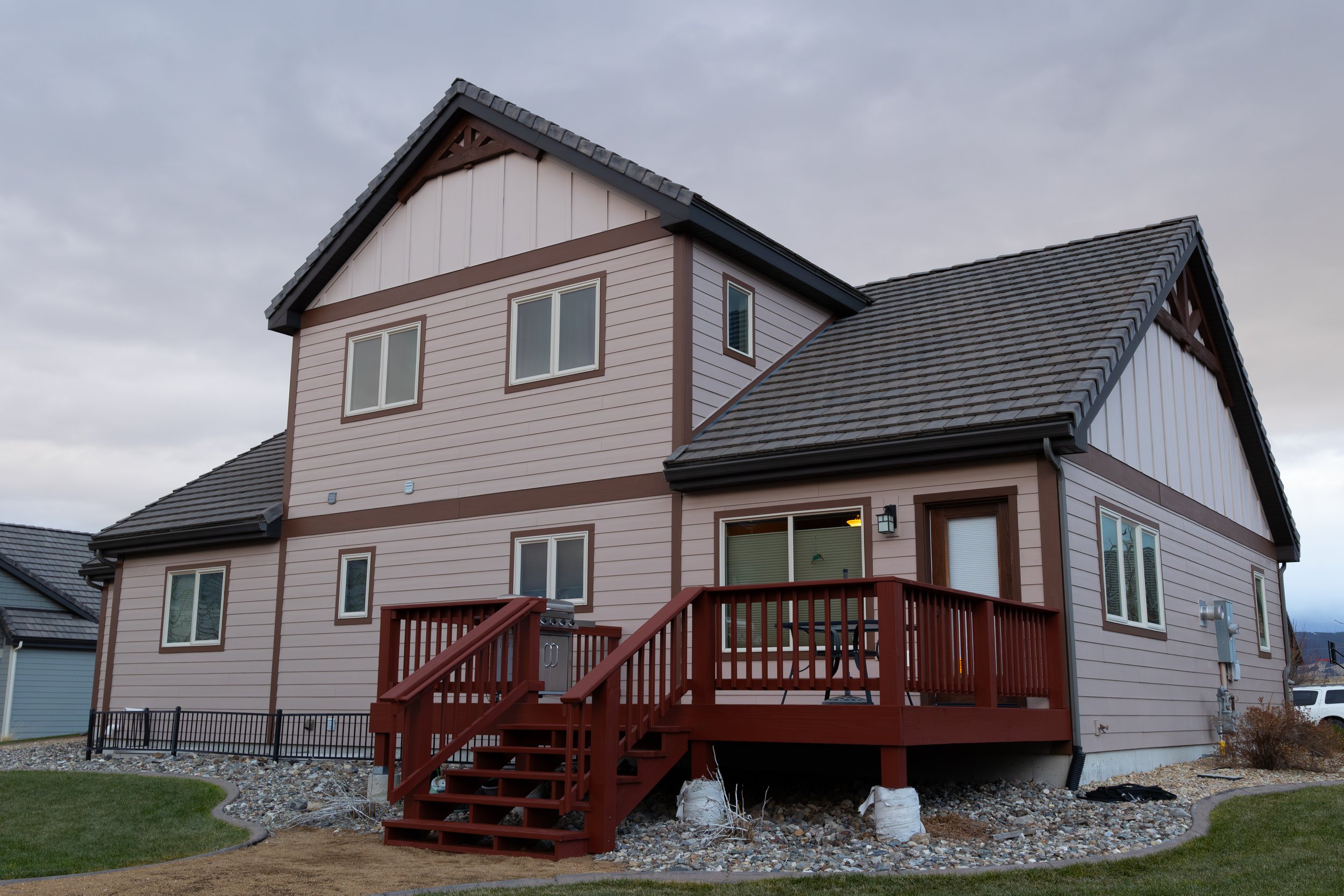 A two-story house with beige siding and brown trim, a small front porch with a red railing, a gray shingles roof, and multiple windows.
