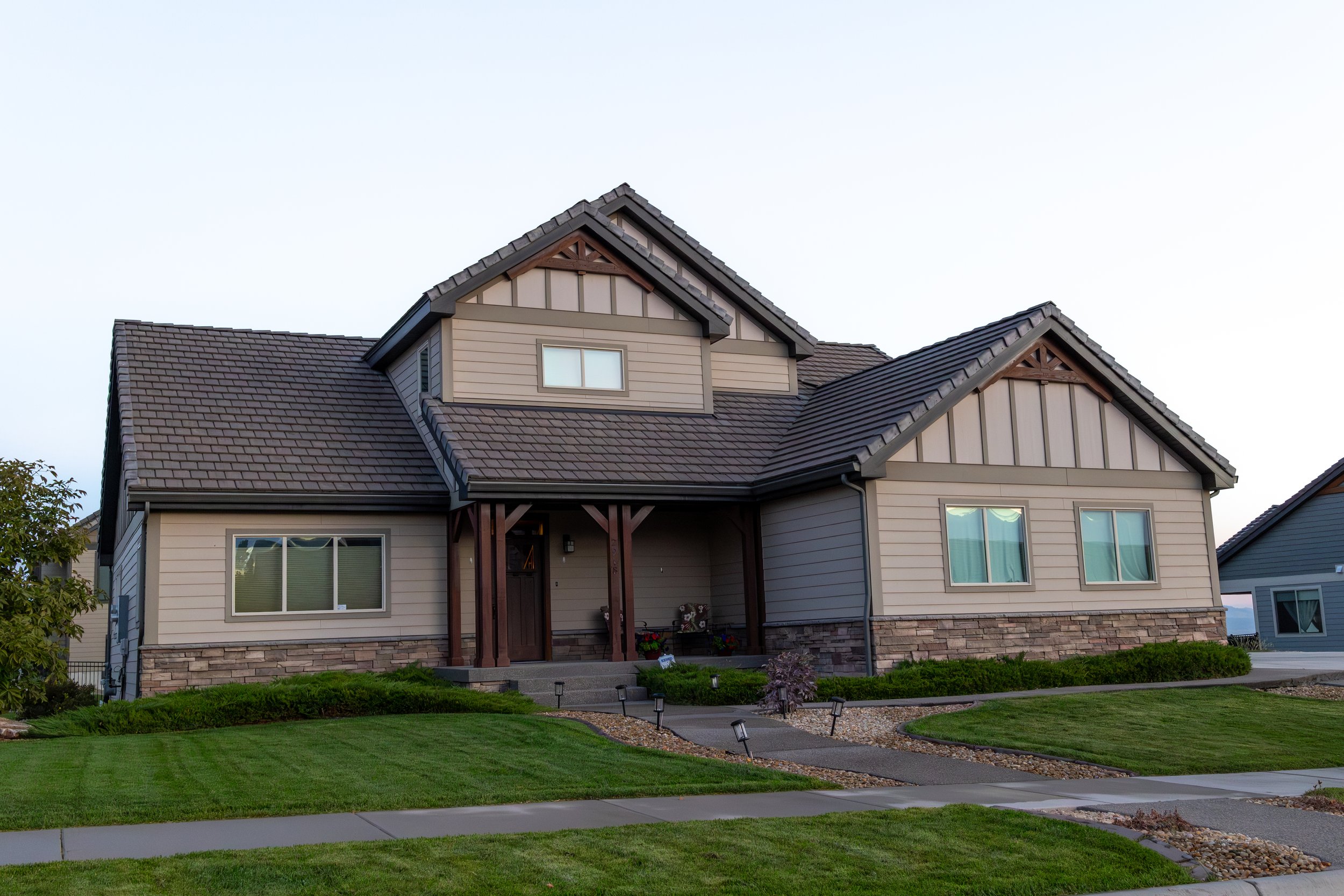A modern two-story house with beige siding, stone accents, and dark gray tiled roof. Front porch with wooden posts, green lawn, sidewalk, and landscaping.