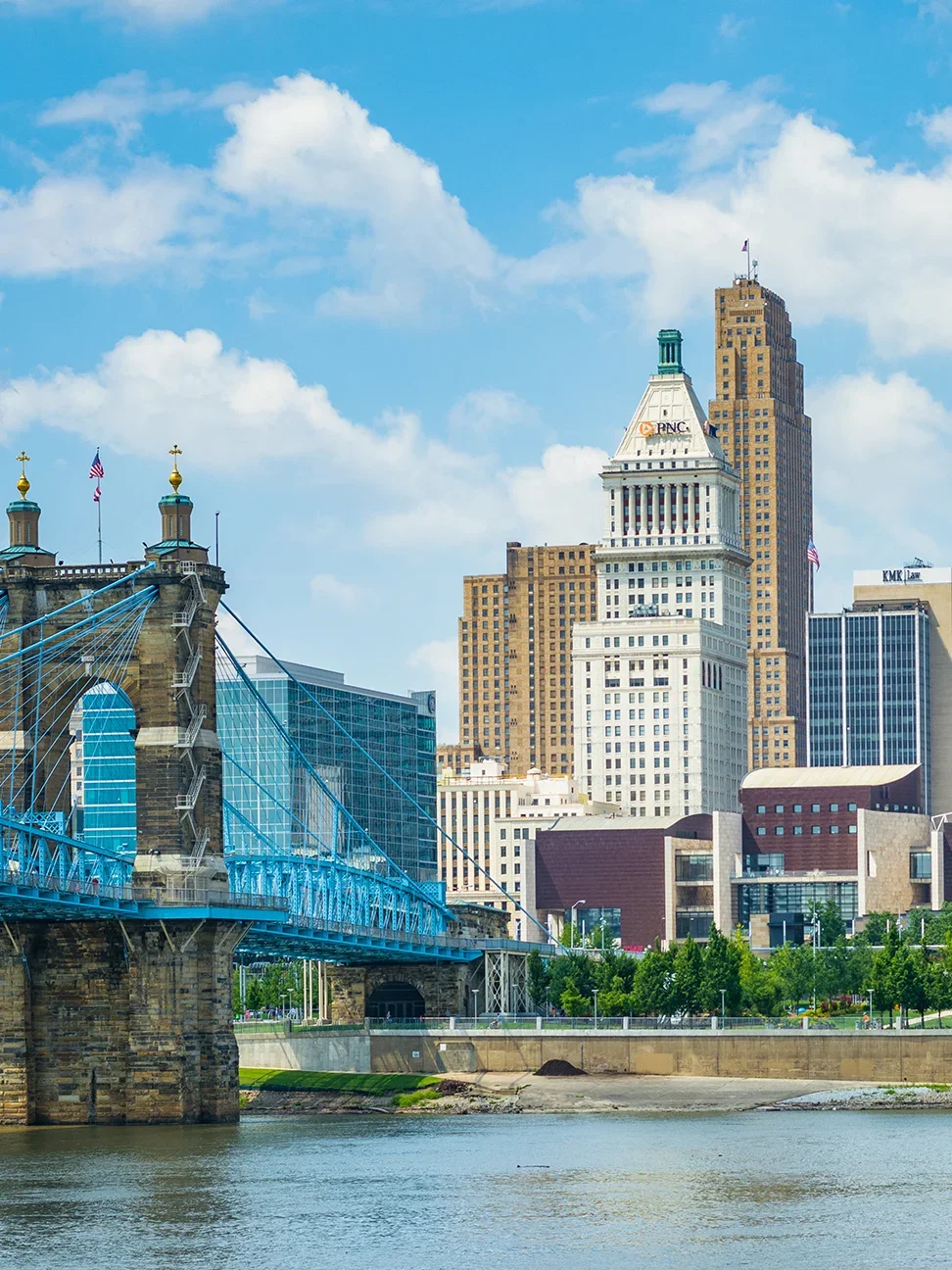 Cincinnati skyline featuring a blue suspension bridge over a river, with tall skyscrapers and a partly cloudy sky in the background.
