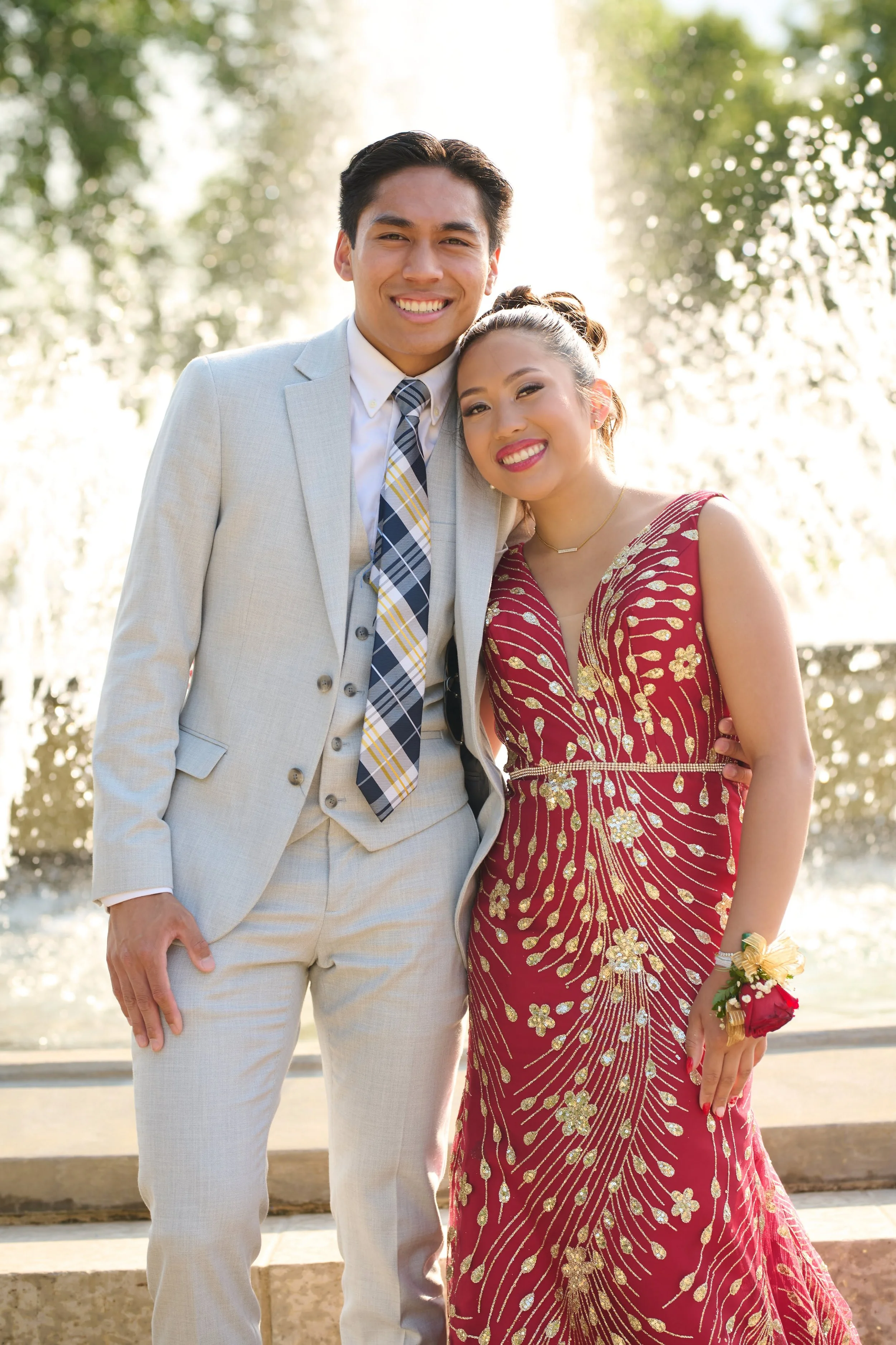A young man in a light gray suit and young woman in a red and gold dress smiling outdoors, standing close together with trees and sunlight in the background.