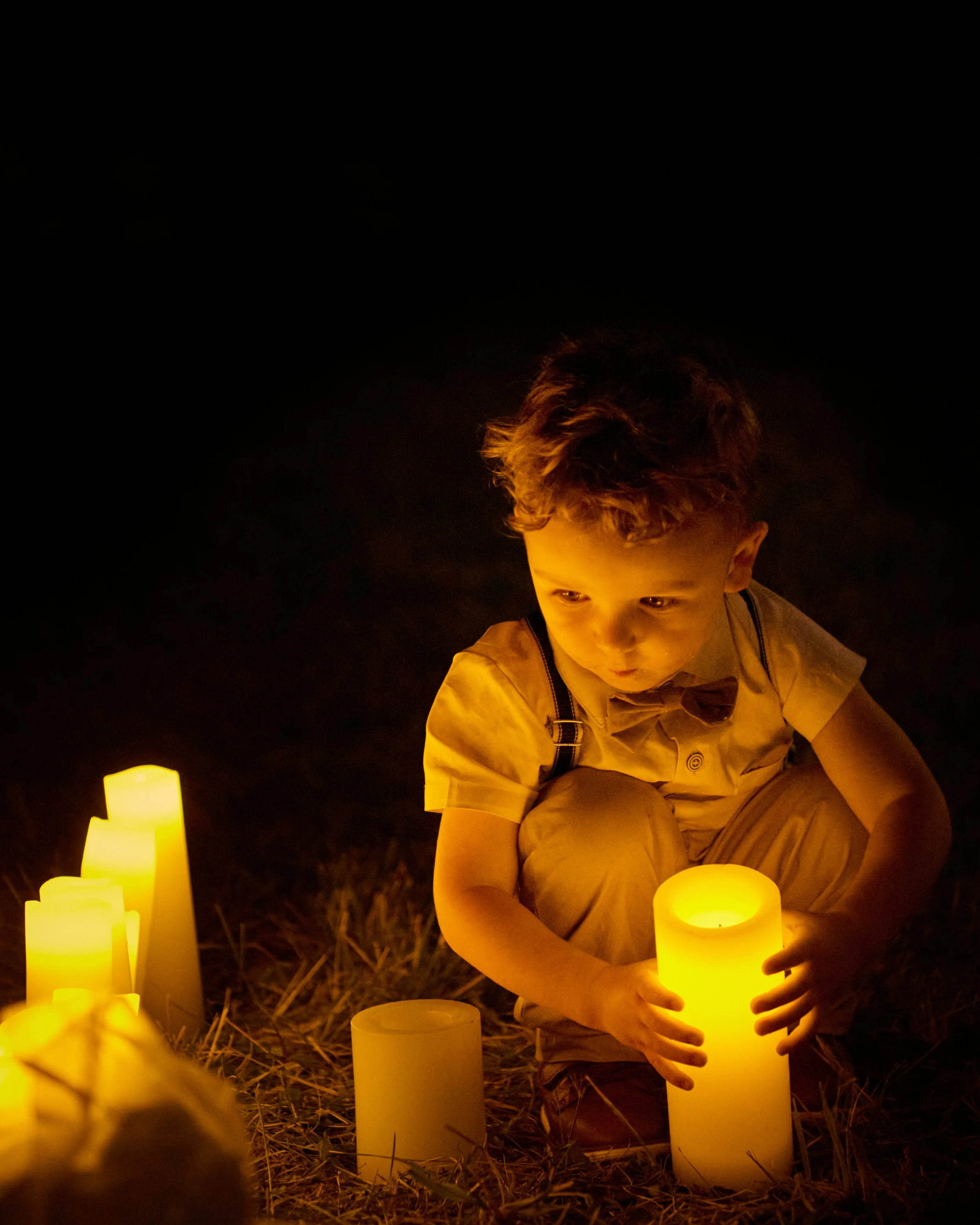 A young boy crouches on the ground at night, holding a lit candle amidst several other lit candles, with a dark background.