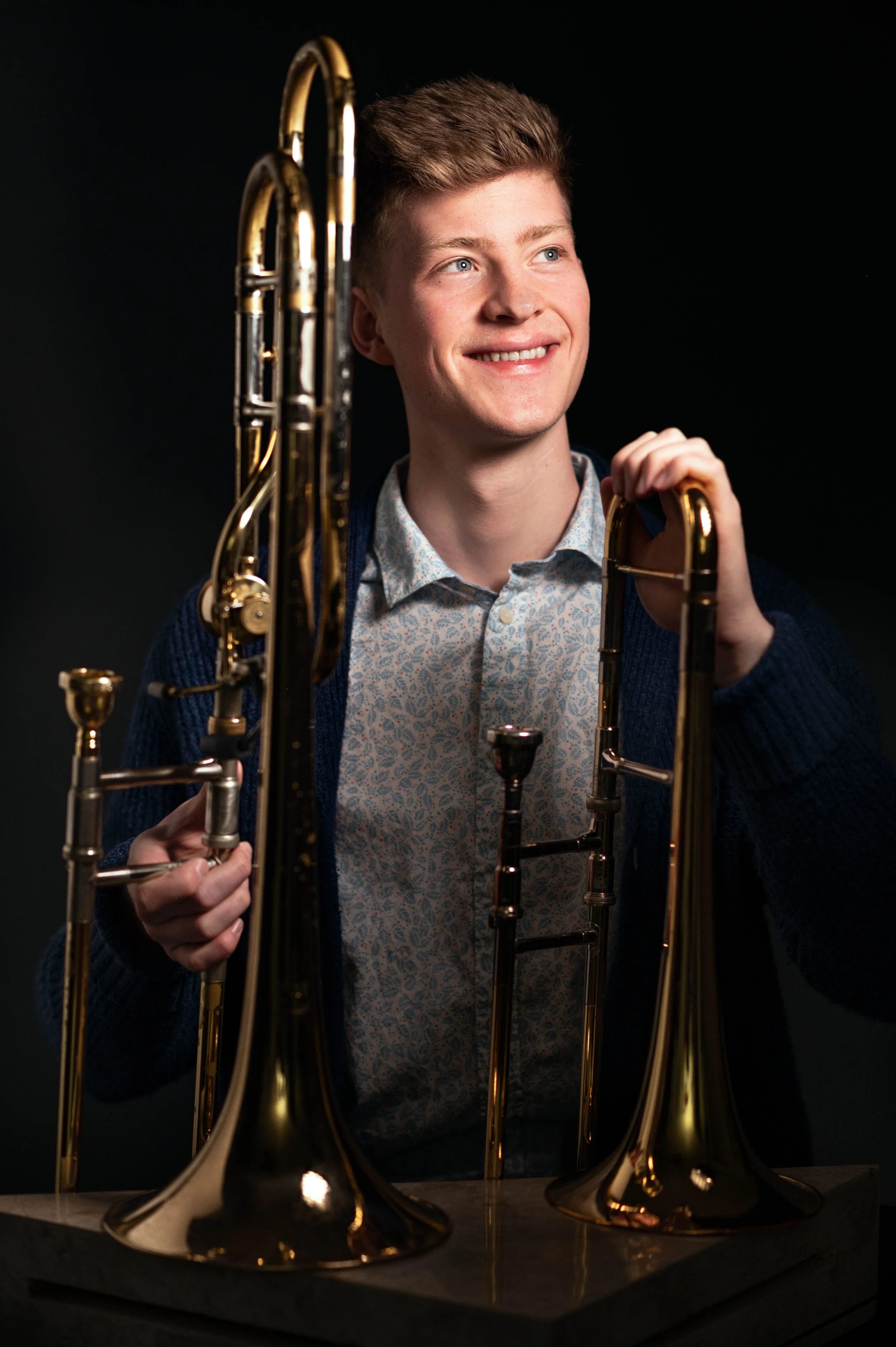 A young man with light brown hair smiling while holding a brass trombone, surrounded by other brass instruments on a dark background.
