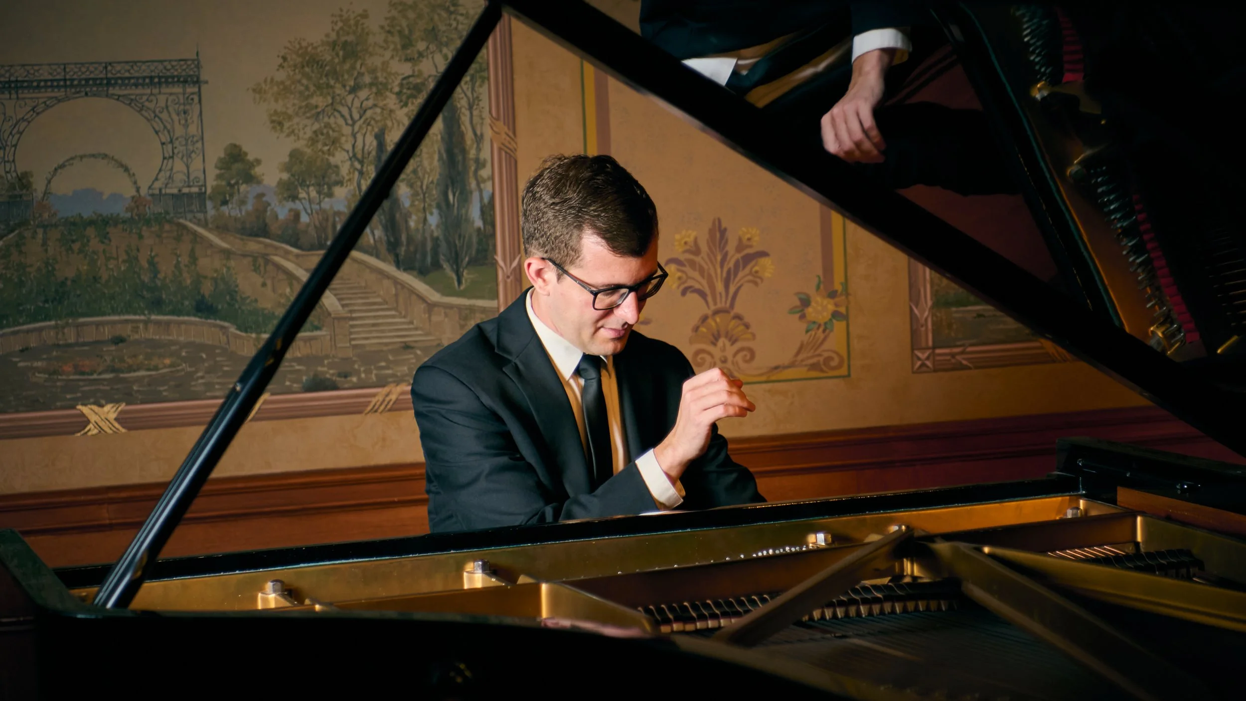A young man wearing glasses, a black suit, white shirt, and a black tie, is sitting at a grand piano, with a scenic mural painting of gardens and trees in the background.
