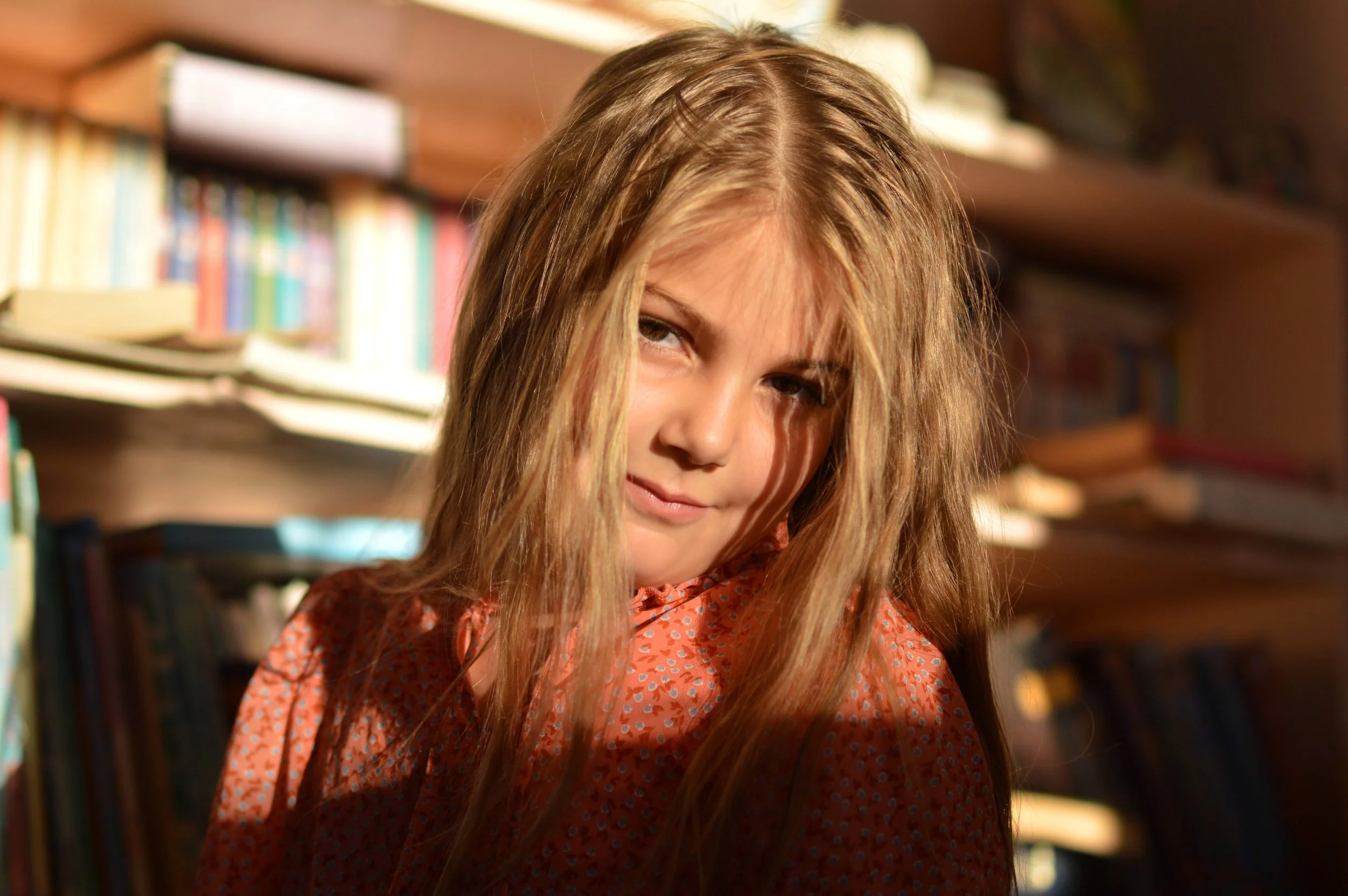A young girl with long, light brown hair and a slight smile, wearing a orange patterned top, in a room filled with bookshelves.
