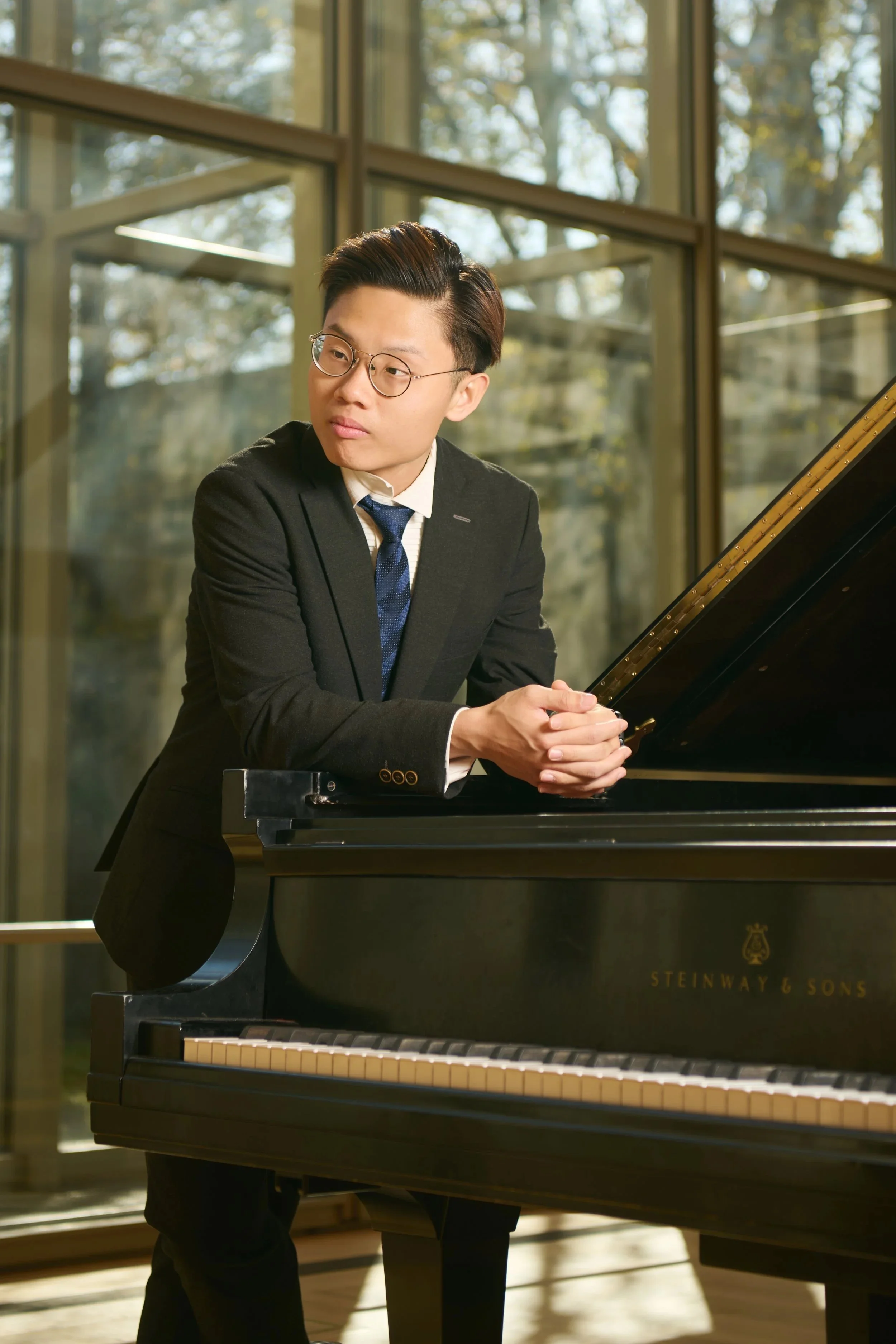 A young man in a black suit and glasses stands next to a Steinway & Sons grand piano in a room with large windows and natural light.