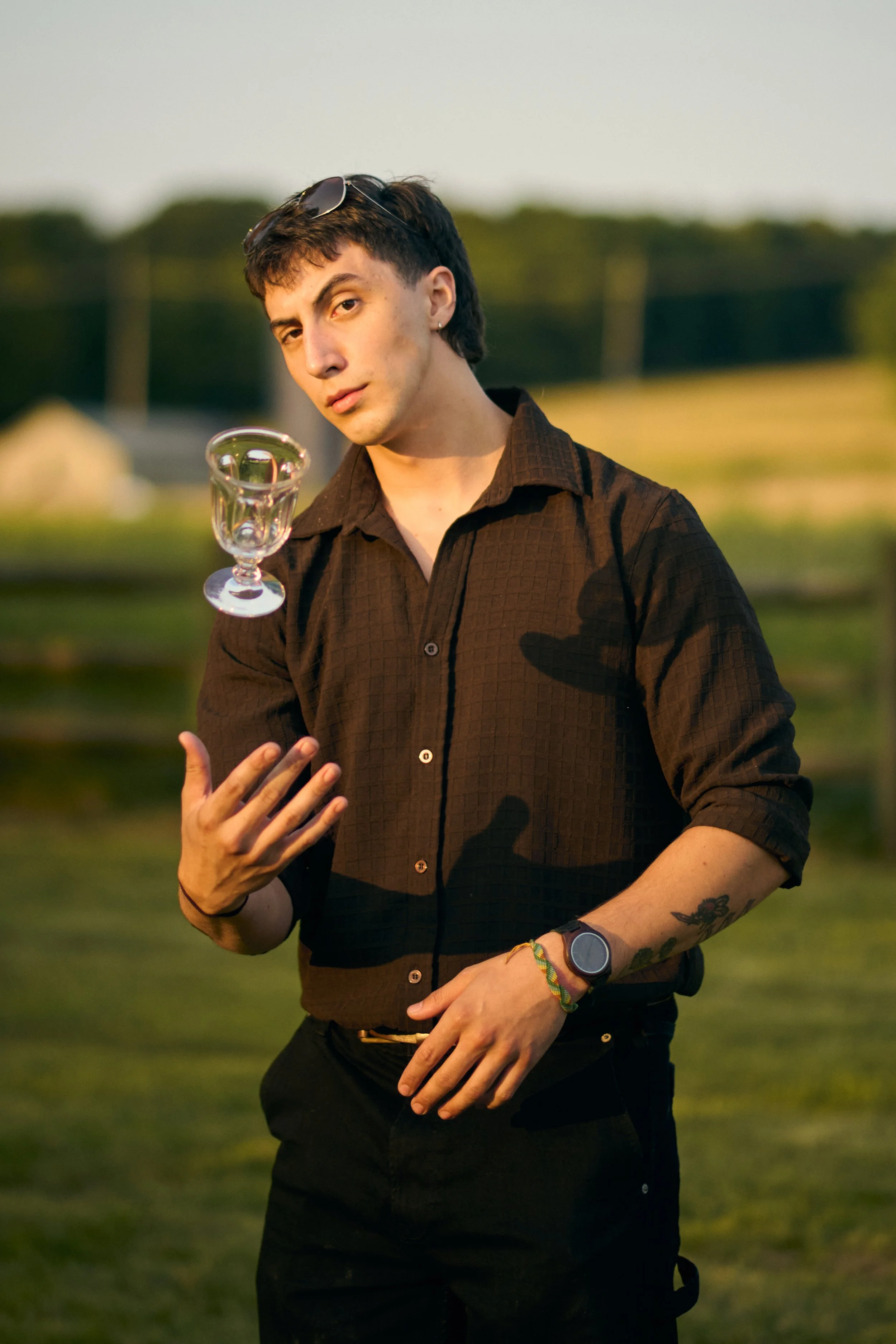 A young person with short dark hair, wearing sunglasses on their head, a black shirt, and a wristwatch, is outdoors in a grassy area during sunset. They are holding a clear glass goblet in one hand and have printed tattoos on their forearm, with a serious expression.