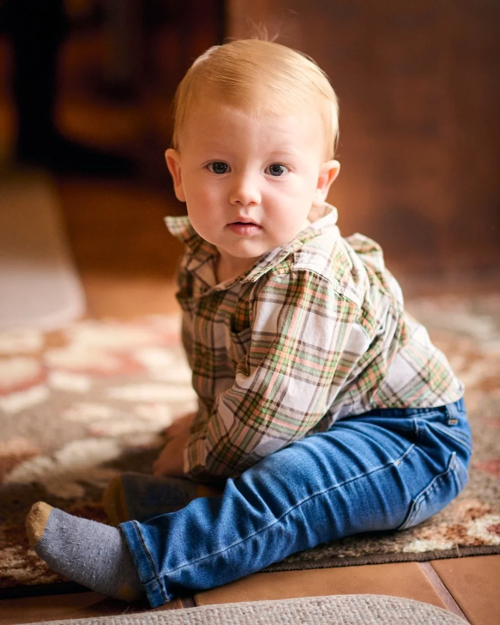 A young boy with light blonde hair and blue eyes is sitting on a patterned carpet, wearing a plaid shirt, blue jeans, and gray socks, looking toward the camera.