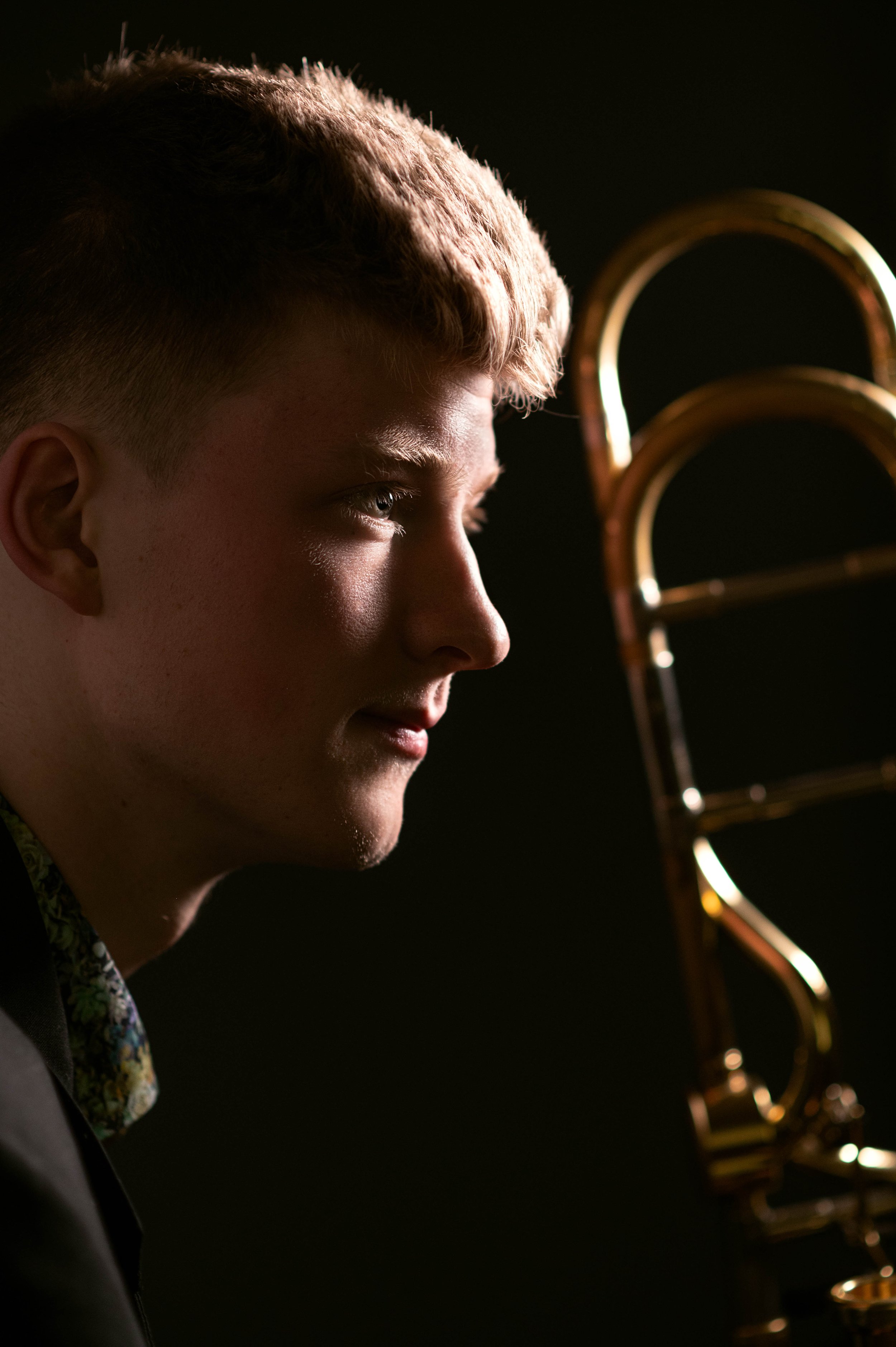 A young man with blond hair is facing left, illuminated by a soft light, with a golden trombone partially visible on the right.
