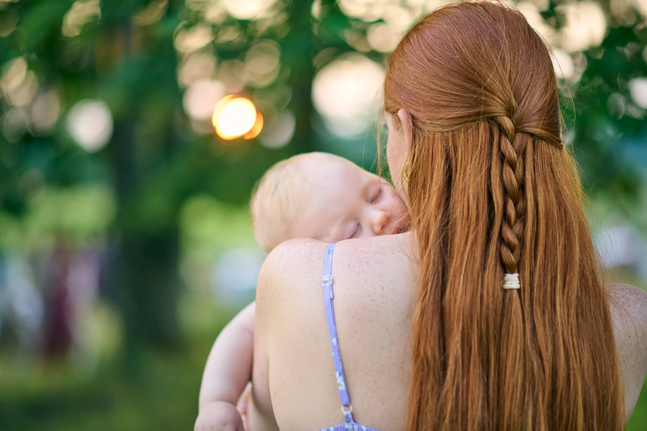 A woman with long red hair styled in a braid holding a sleeping child with blond hair outdoors with green trees and sunlight in the background.