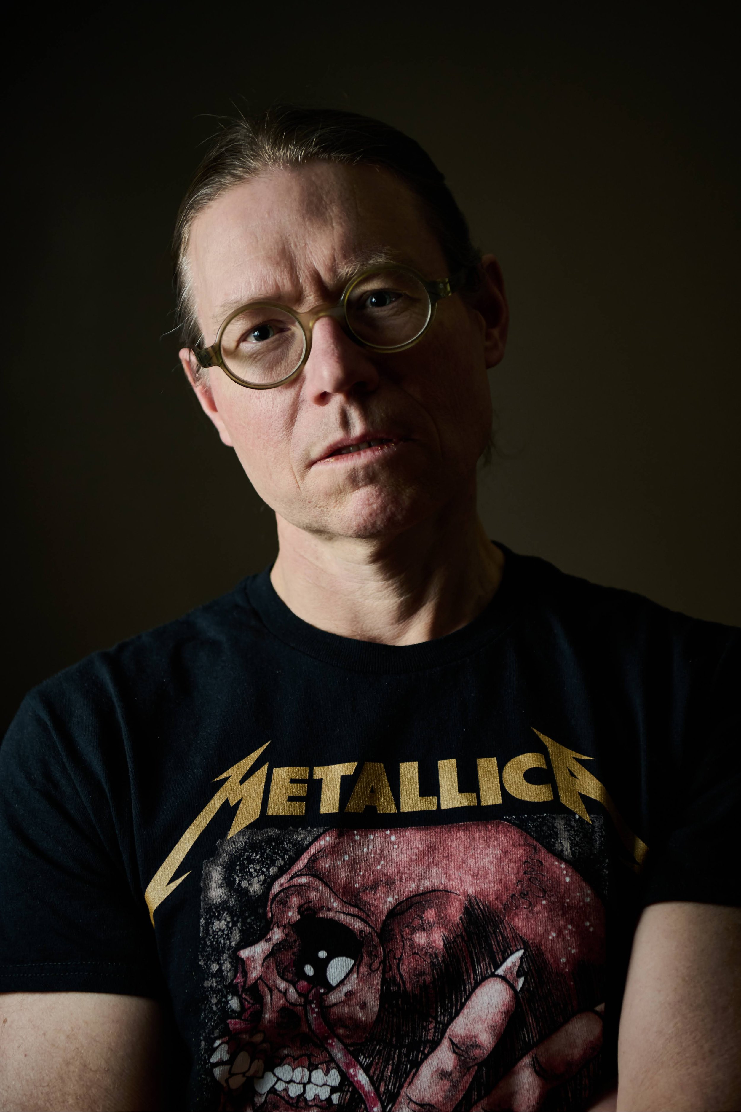 A man with glasses and a Metallica T-shirt takes a close-up selfie with a dark background.
