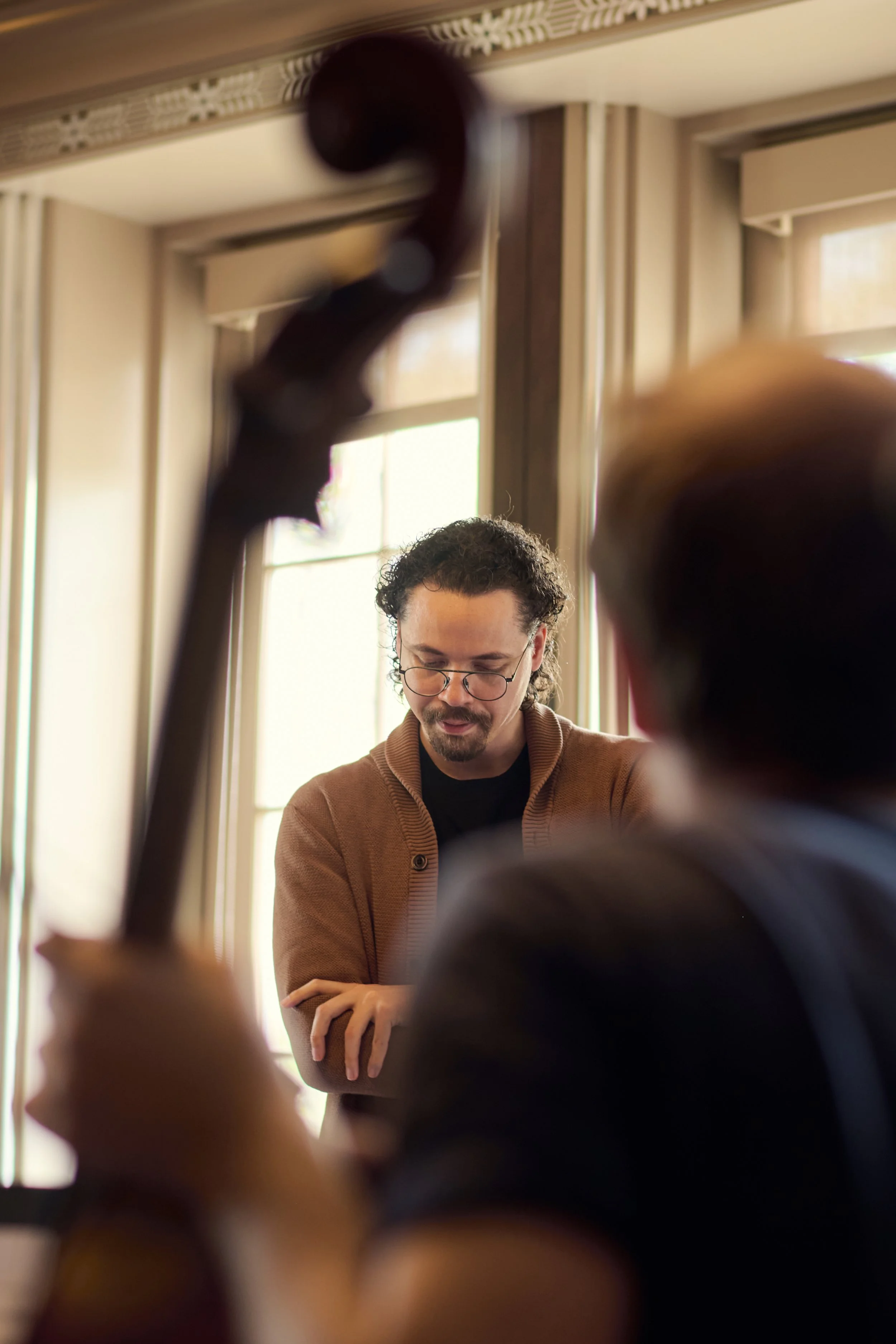 A person with glasses and a brown jacket standing near a piano. Two other individuals are partially visible in the foreground, with one holding a cello. The setting appears to be a well-lit room with large windows.