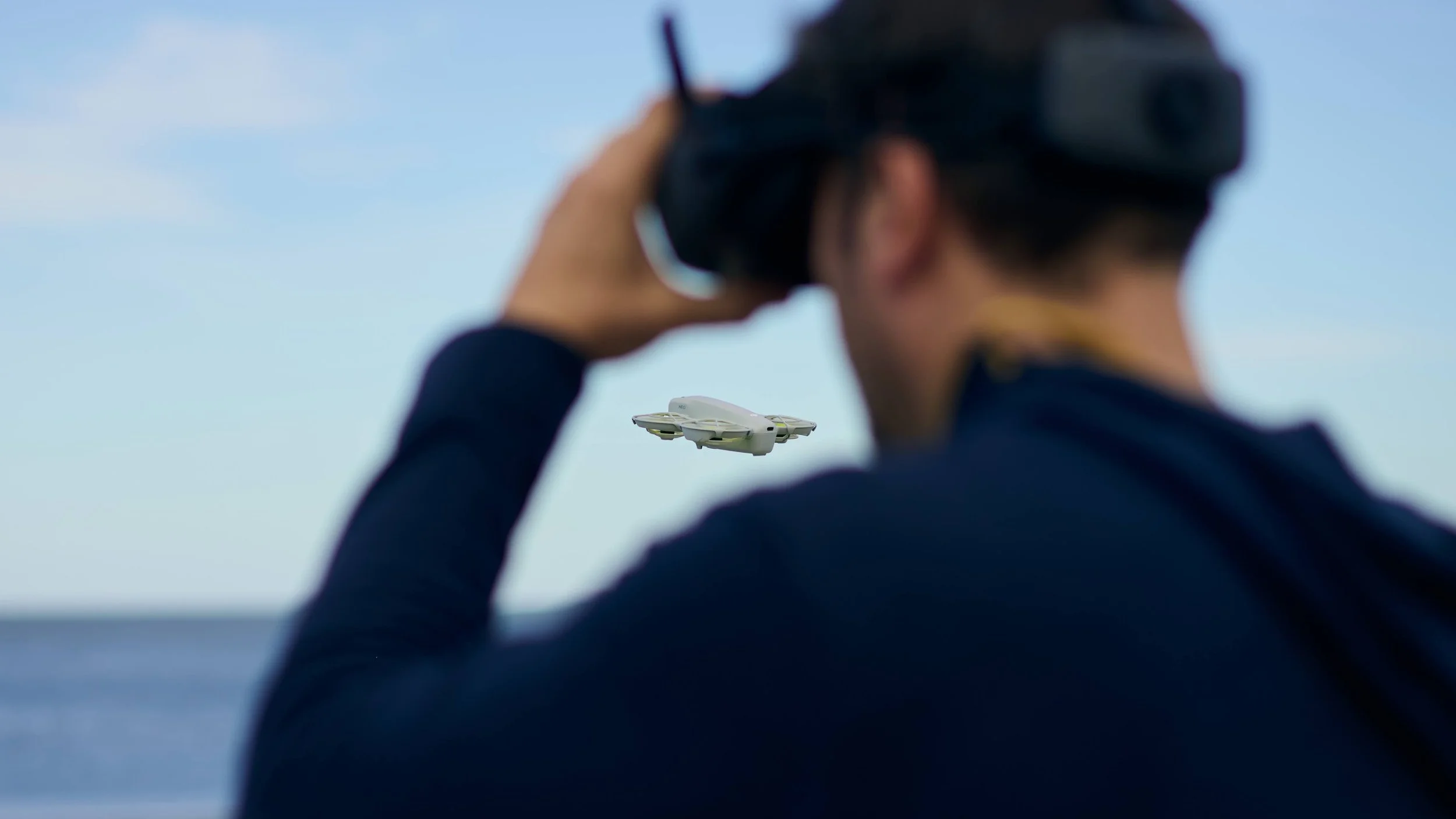 A person wearing a virtual reality headset and a dark long-sleeve shirt, holding a remote control to their ear, as a small drone flies in the background against a sky and water scene.
