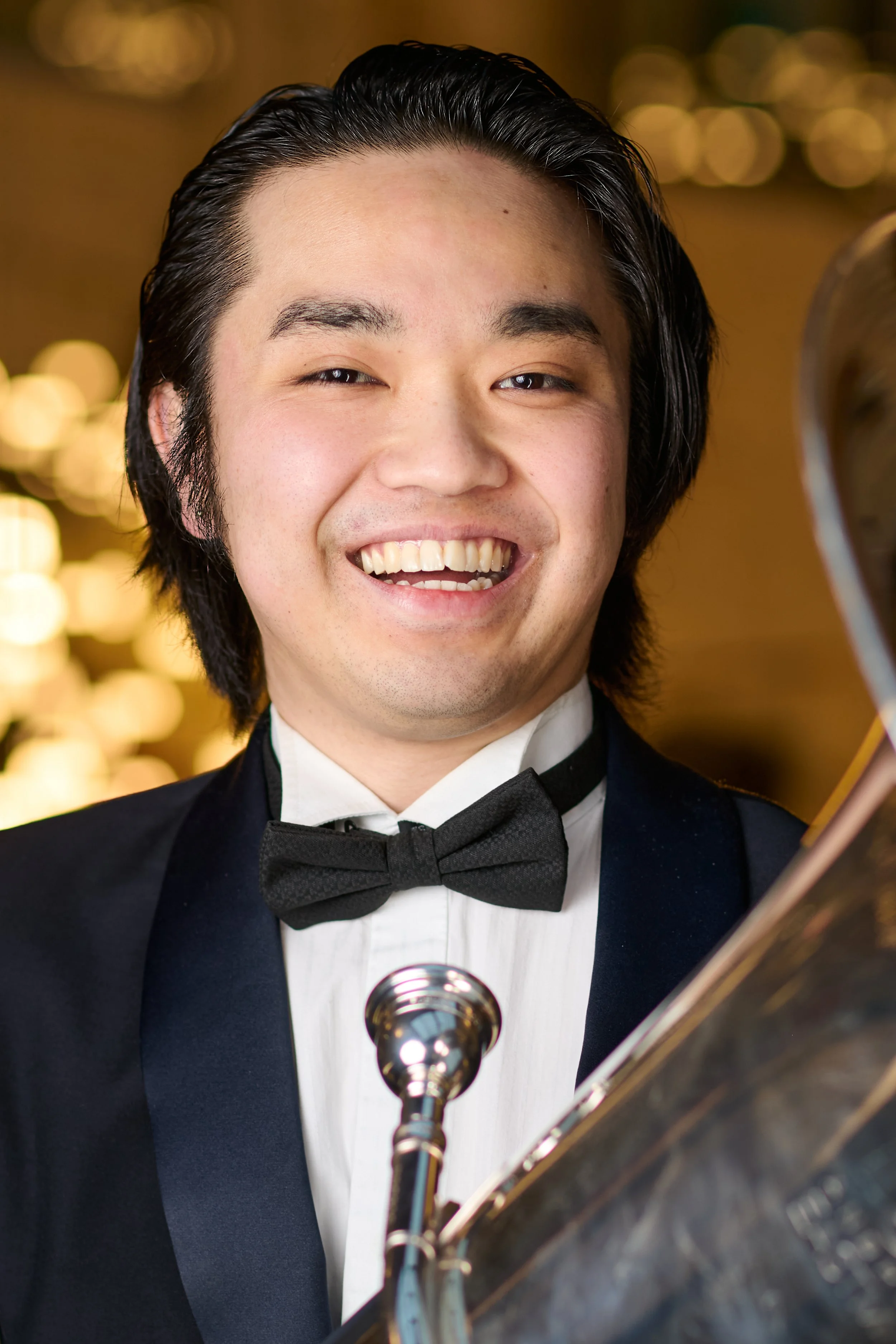 A man with medium-length black hair, wearing a tuxedo with a black bow tie, smiling and holding a tuba in a warmly lit setting.