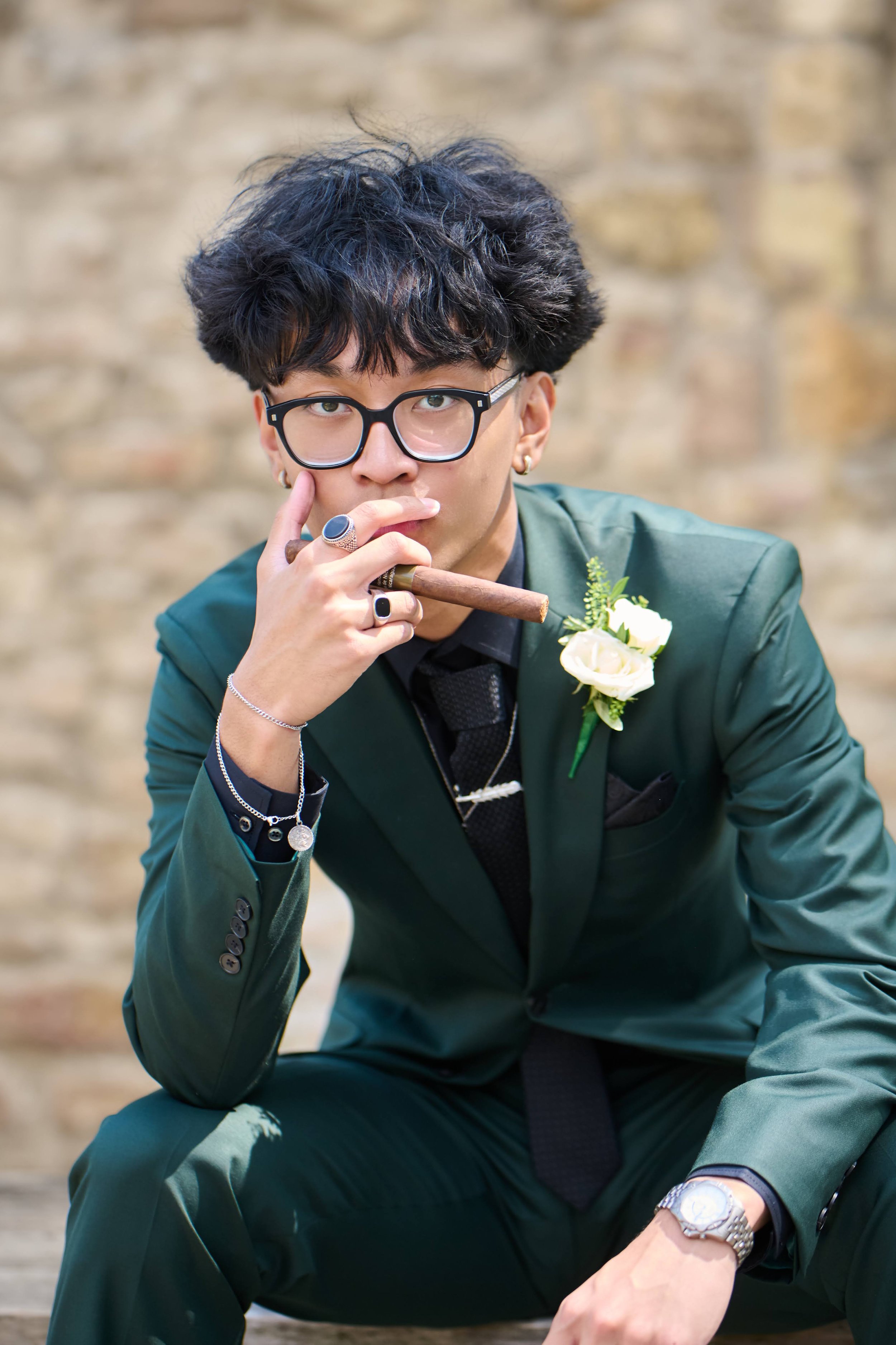 A young man with glasses, dark curly hair, and accessories, wearing a green suit with a white flower boutonniere, sitting outdoors against a brick wall, holding a cigar in one hand while touching his lips with fingers, and looking directly at the camera.