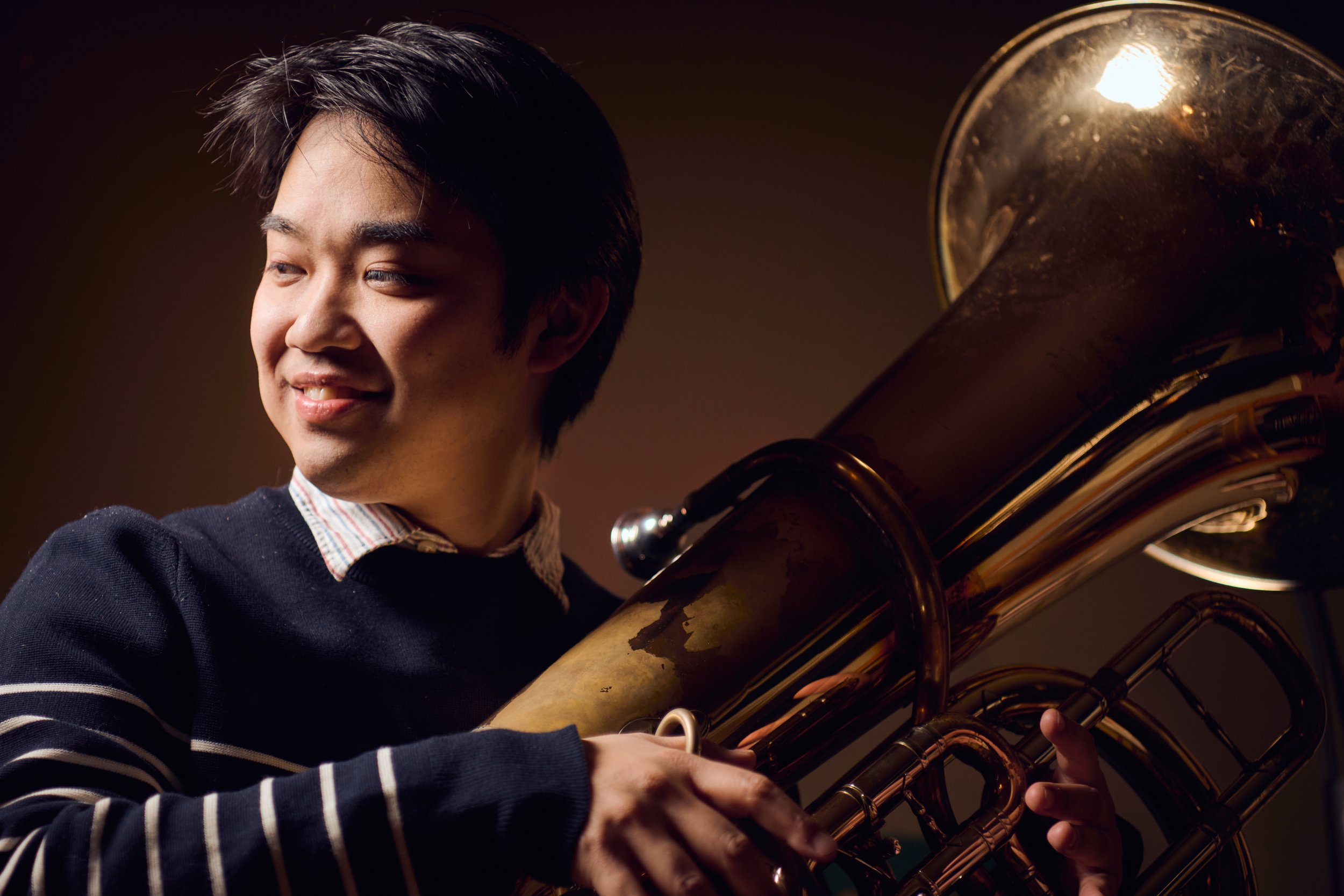 A young man holding a euphonium, smiling and looking to the side, with a dark background.