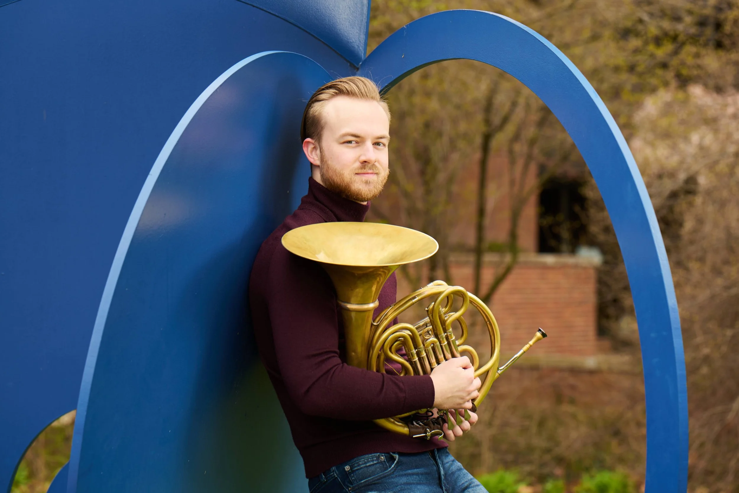A man with blonde hair and a beard holding a brass French horn and a brass tuba, standing outdoors next to a large blue curved metal structure, with trees and a brick building in the background.