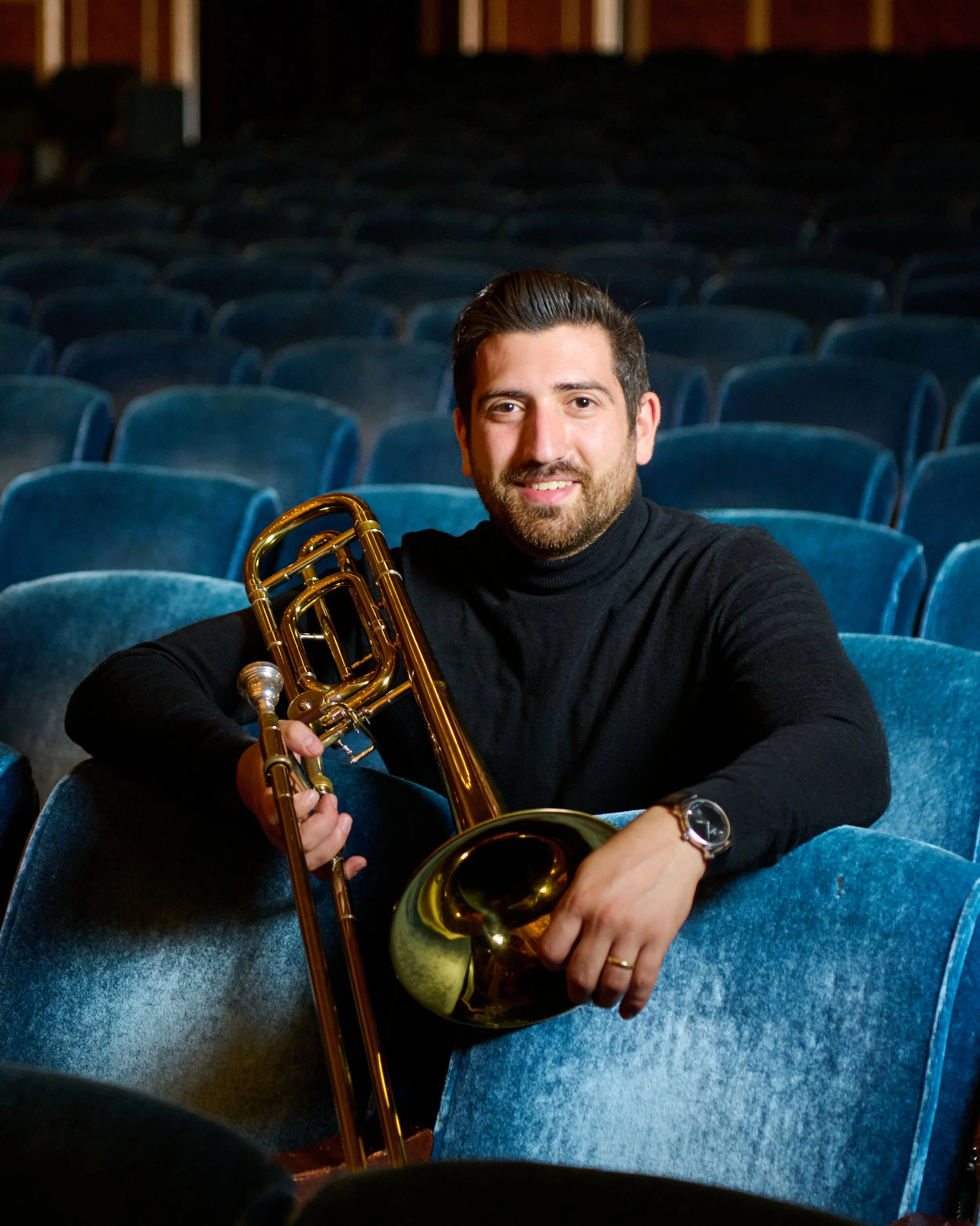 A man with dark hair and a beard sitting in an empty auditorium with blue seats, holding a brass trombone, smiling at the camera.
