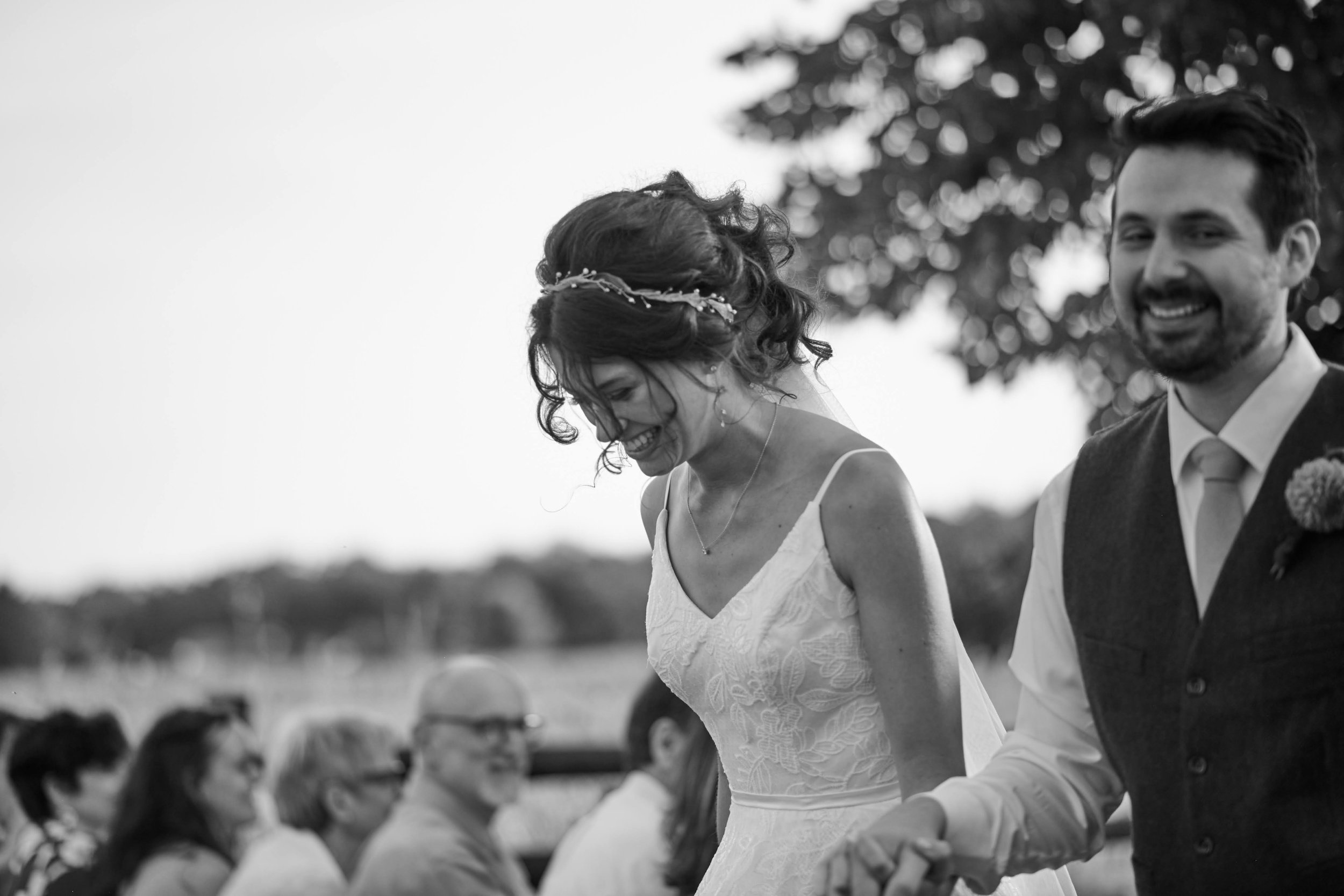 Black and white photo of a bride and groom holding hands, smiling during their outdoor wedding ceremony, with guests seated in the background.