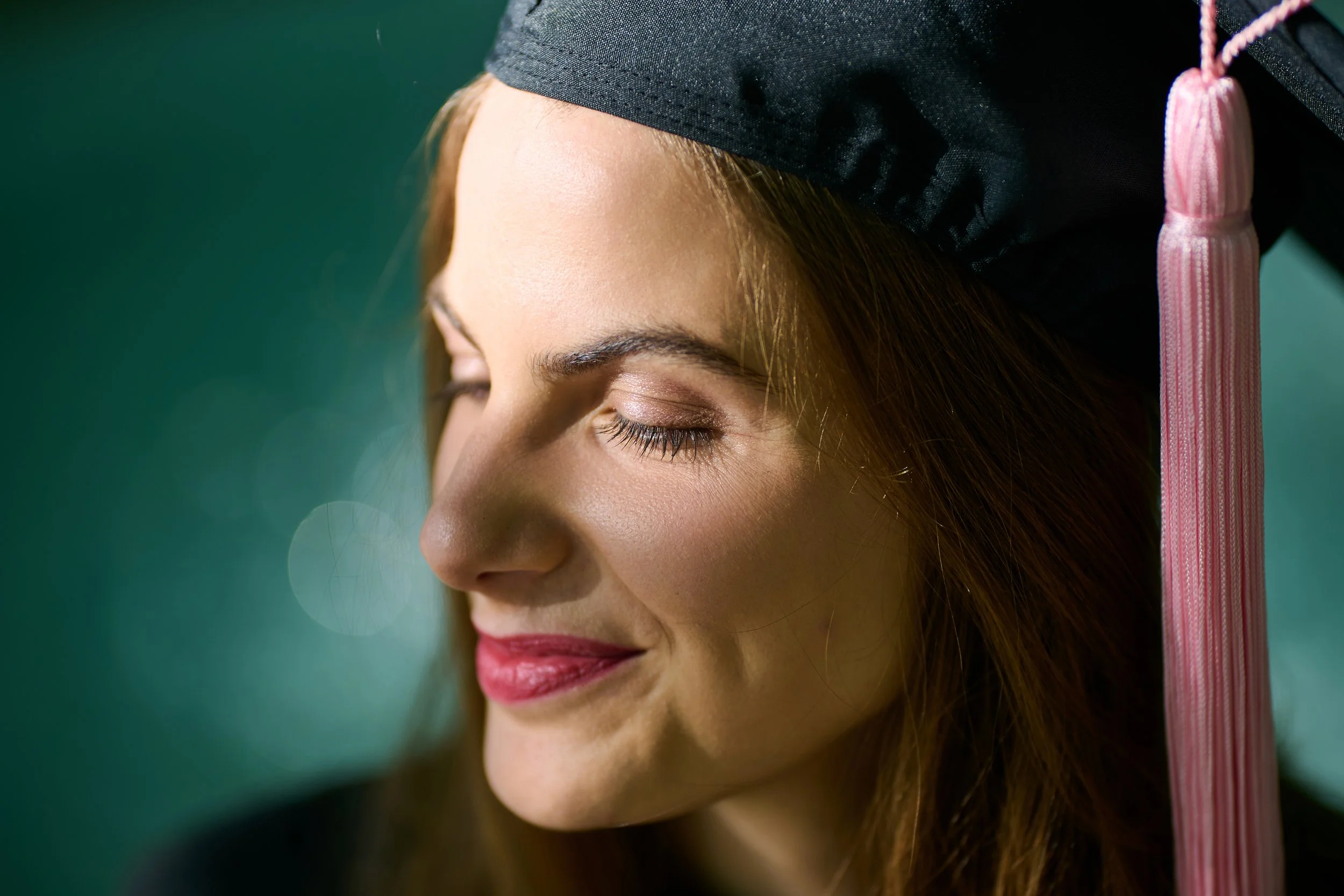 Close-up of a young woman with eyes closed, wearing a graduation cap with a pink tassel, smiling gently.