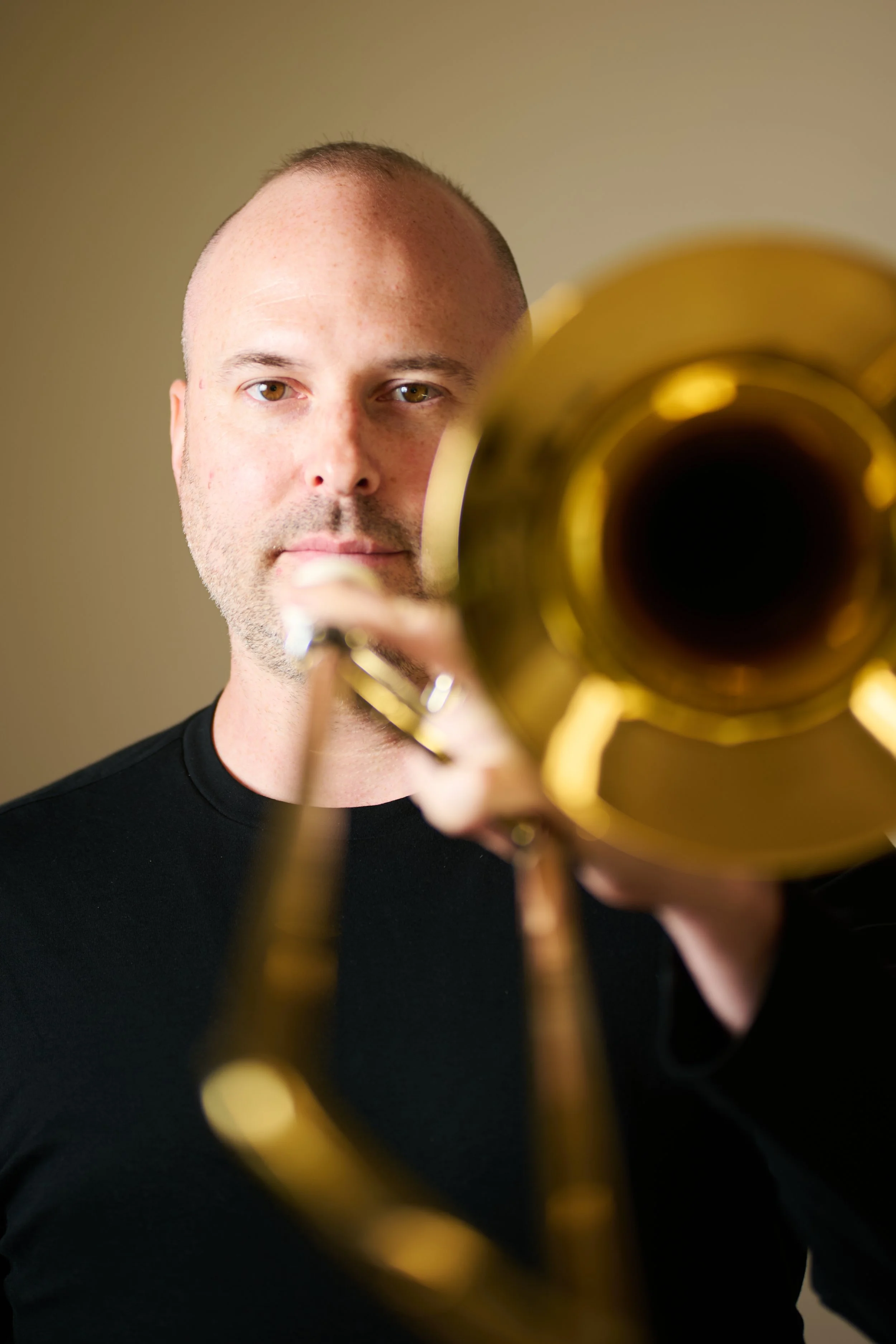 A man with short hair and wearing a black shirt, holding a brass trumpet towards the camera, with the trumpet's bell close to the lens.
