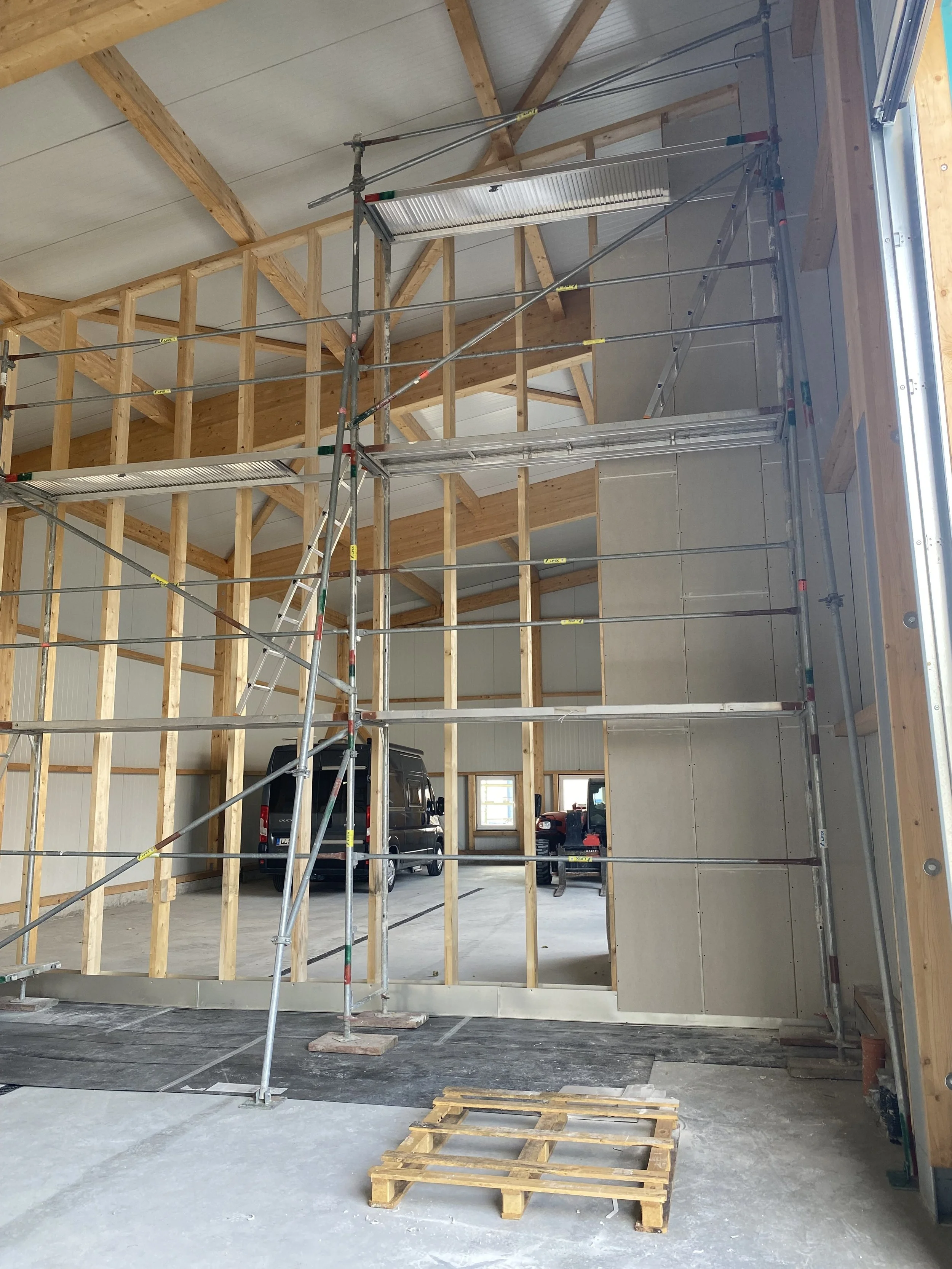 Innenansicht einer Baustelle mit Holzrahmen, Gerüst und geparkten Fahrzeugen im Hintergrund.