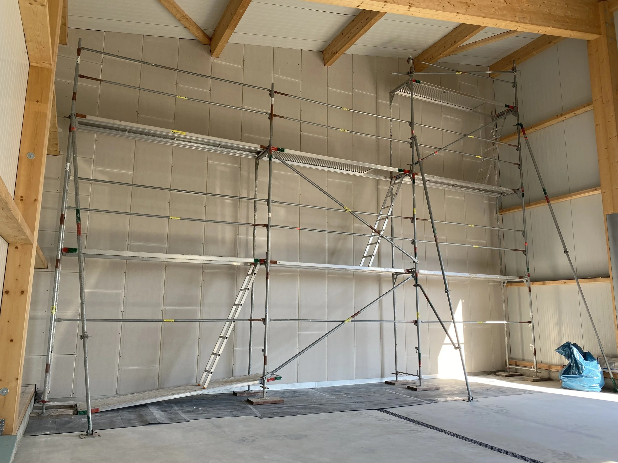 Baugerüst in einem Raum unter einem Dach mit Holzdecken, noch unfertige Wände, eine Leiter, und ein blauer Müllsack auf dem Boden.