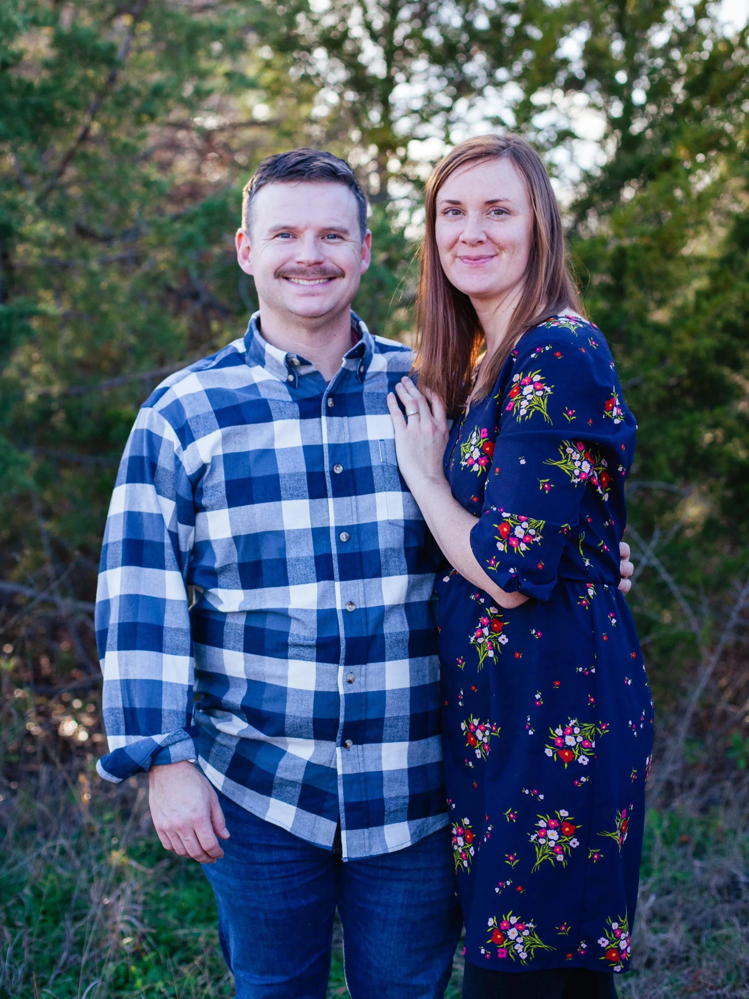 A man and a woman standing outdoors in front of trees, smiling at the camera. The man is wearing a blue and white plaid shirt, and the woman is in a dark blue dress with floral patterns.