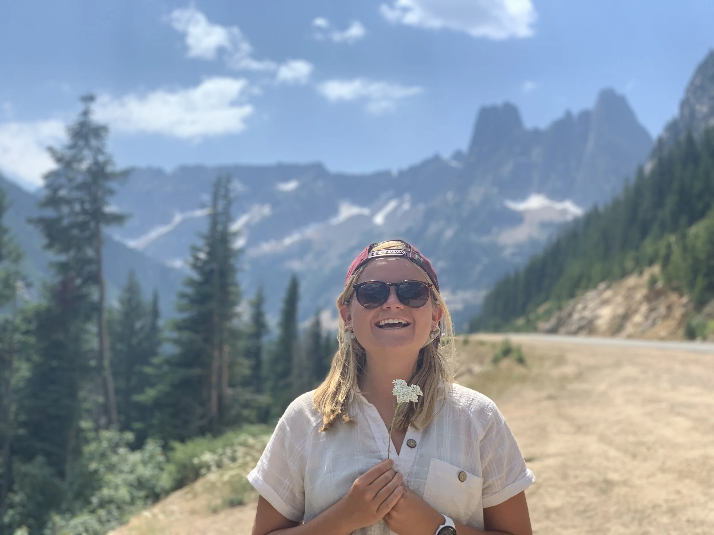 A woman wearing sunglasses and a baseball cap smiles while holding a small white flower, with mountains and evergreen trees in the background on a sunny day.