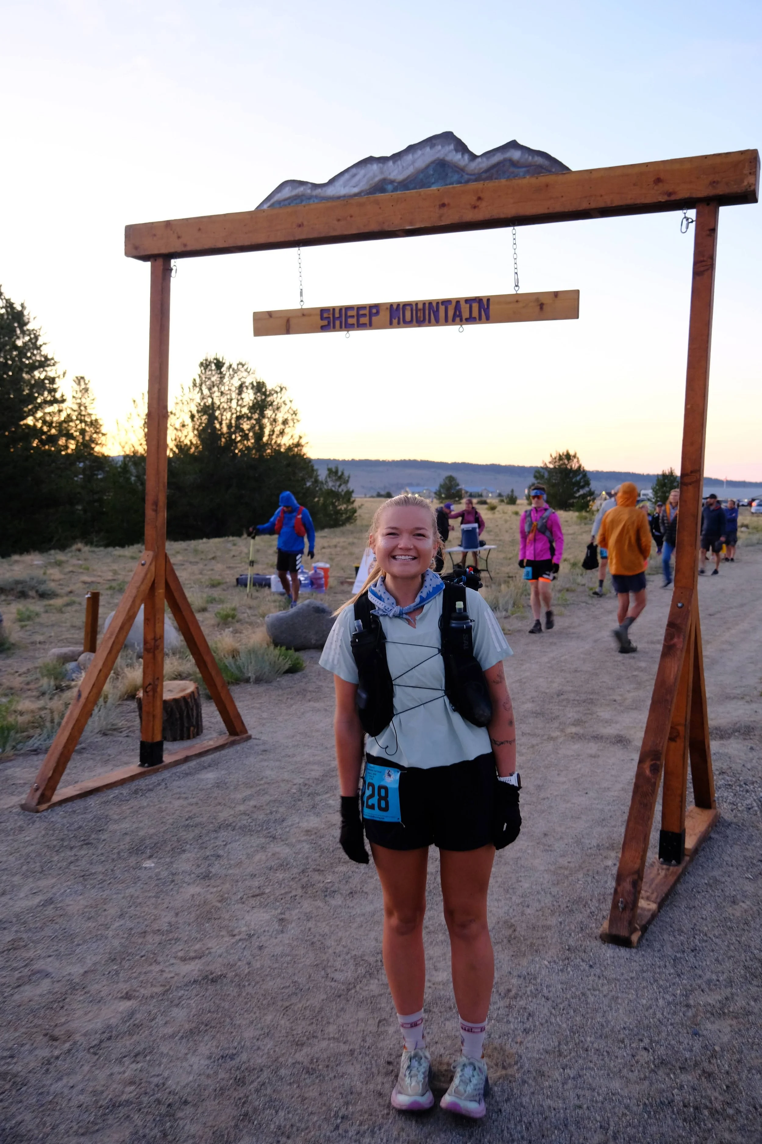 Smiling woman standing at the start of a race at Sheep Mountain, with a wooden archway and distant mountain in the background.