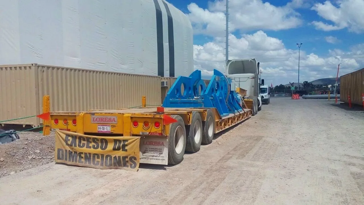 Camión de plataforma con señal de aviso que dice 'Acceso de dimensiones', en un área de construcción con contenedores y estructuras temporales.