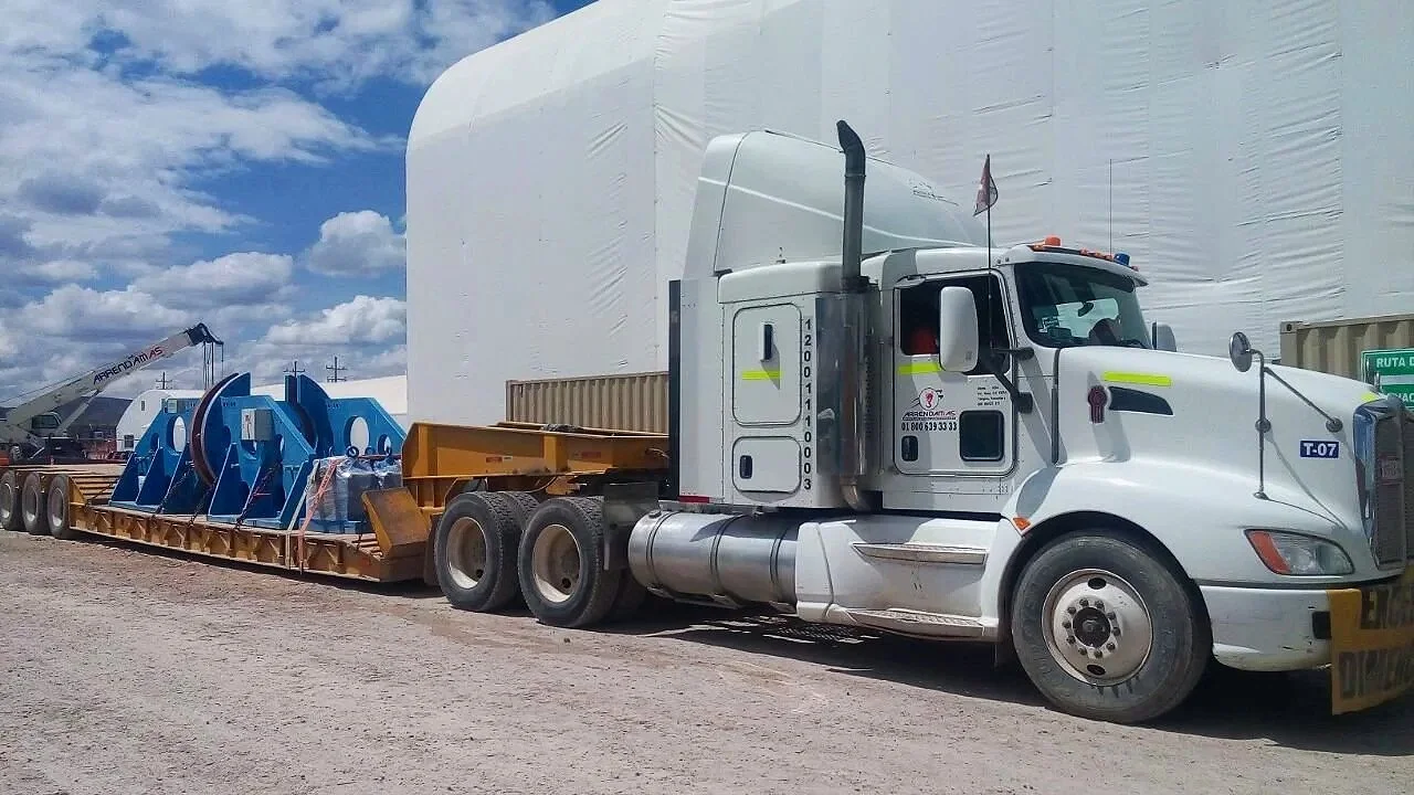 Camión de carga amarillo transportando maquinaria azul en una carretera con cielo nublado.