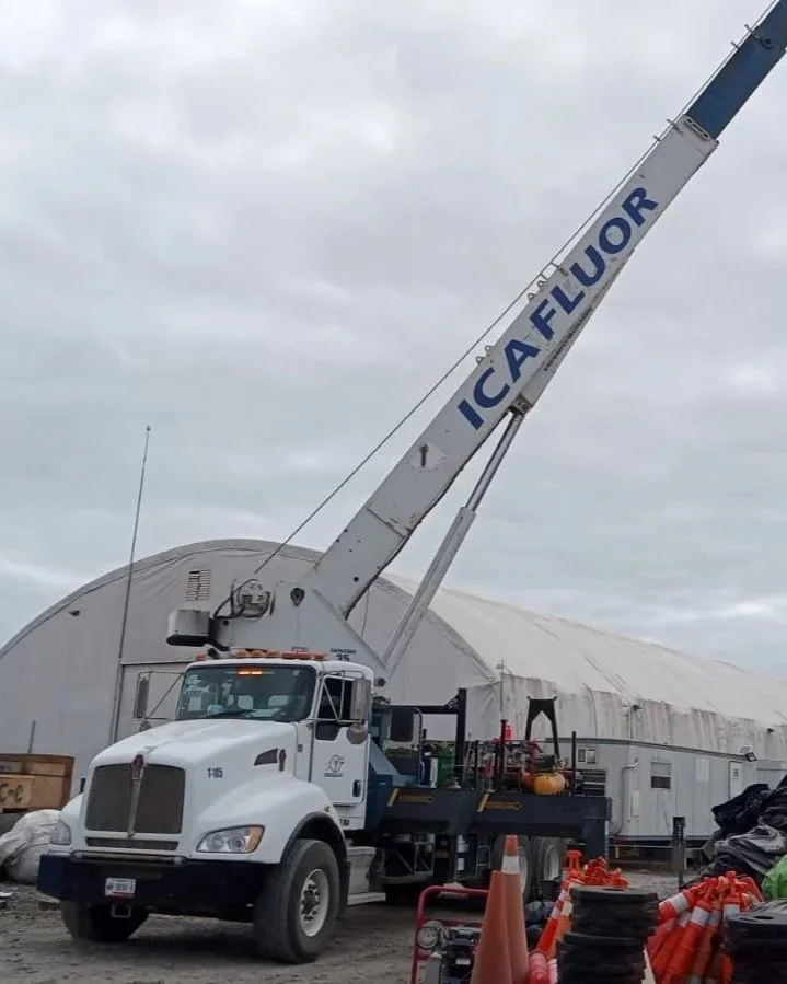 Una grúa con la palabra 'ICALIFLUOR' en la pluma, estacionada frente a un Quonset en un área de construcción con conos naranjas y algunos materiales de construcción alrededor.