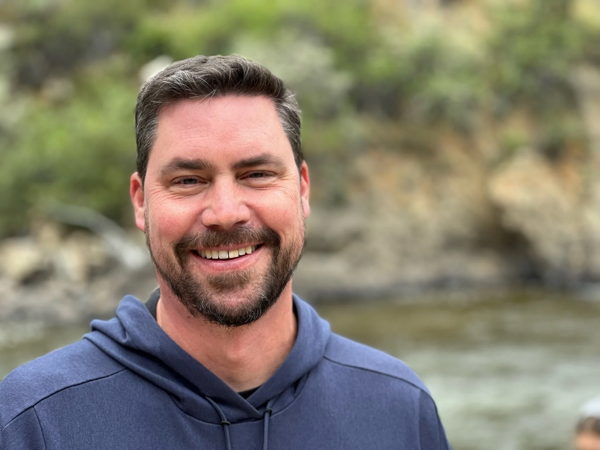 Headshot of writer Ben Fogelberg, smiling with blurred bank of the Poudre River in the background.