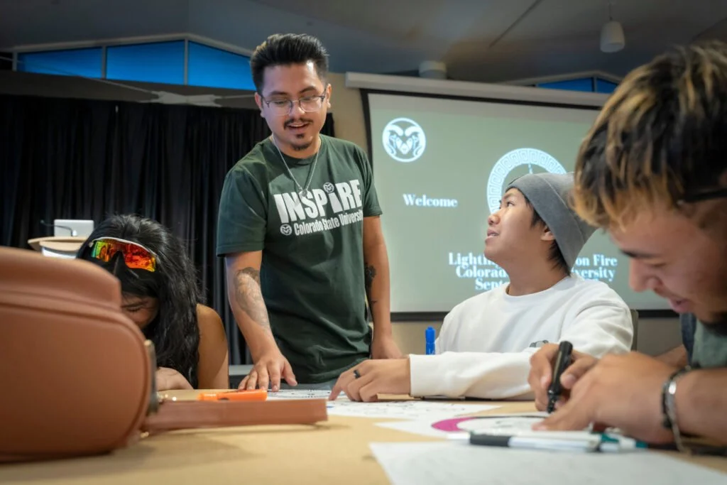 A man standing and talking to three seated young adults in a conference room, with a presentation slide in the background that reads 'Welcome.' The group is engaged in a discussion or activity at a table with notebooks and writing utensils.