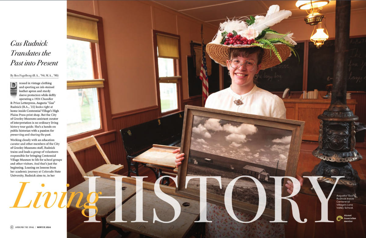 A woman wearing a large straw hat decorated with flowers and berries, smiling and holding a framed black and white landscape photo inside a vintage-style room with wood-paneled walls, an American flag, and chalkboards in the background.