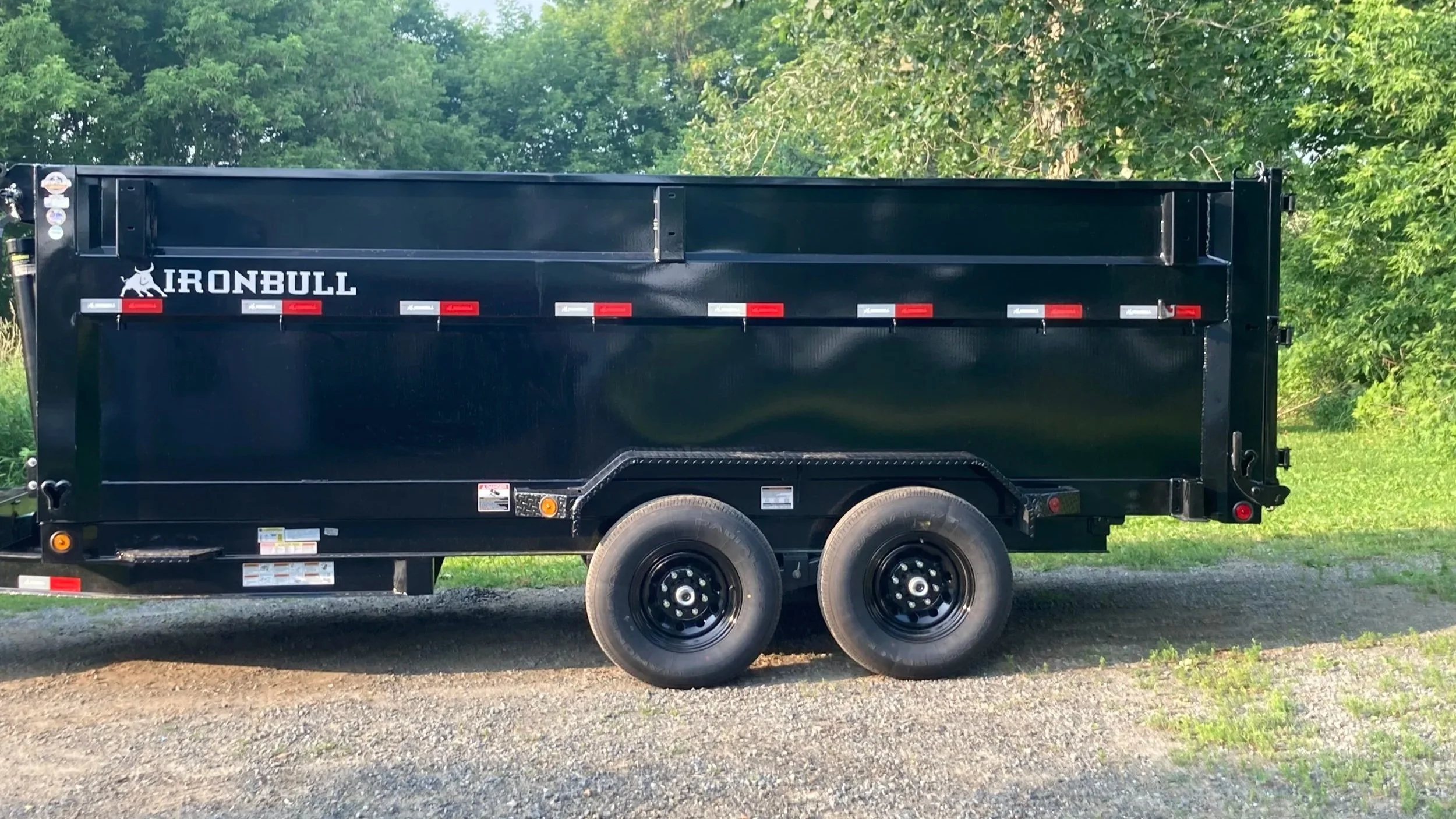 A black utility trailer with the word 'IRONBULL' on its side, parked outdoors on a gravel surface with green trees and bushes in the background.