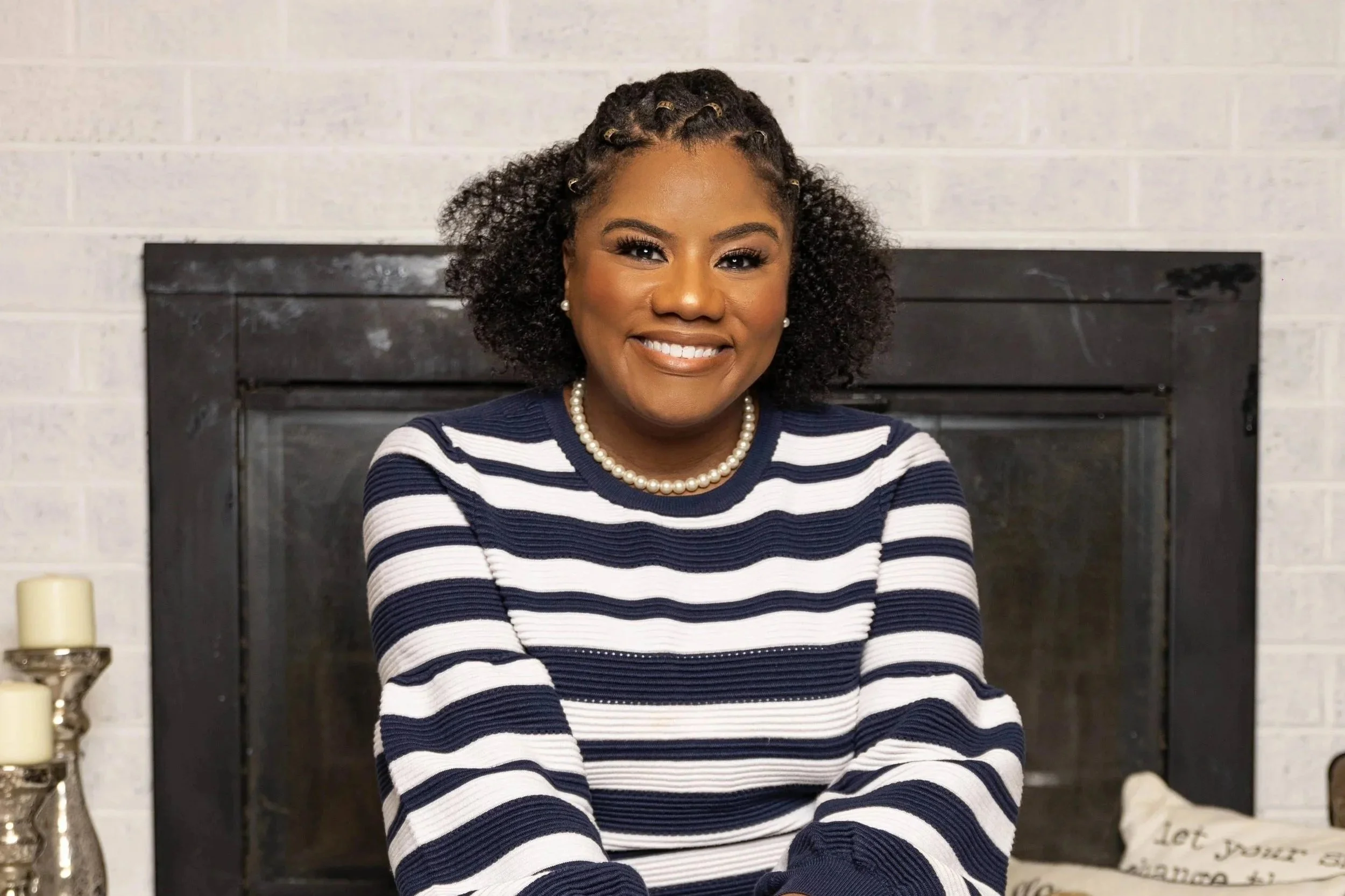 A smiling woman with curly hair, wearing a navy and white striped sweater, pearl necklace, and earrings, sitting in front of a fireplace with a light-colored brick wall in the background.