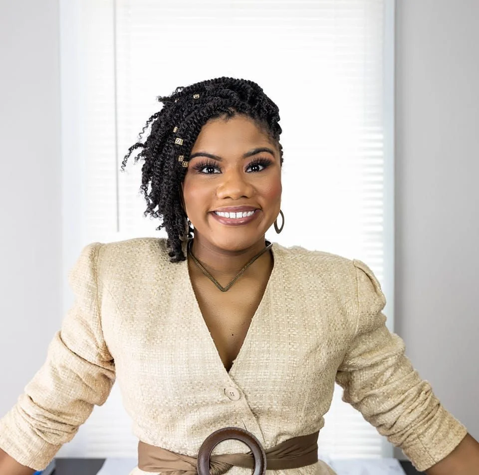 A woman with styled curly hair, wearing a beige blazer and jewelry, smiling in front of a window with blinds.