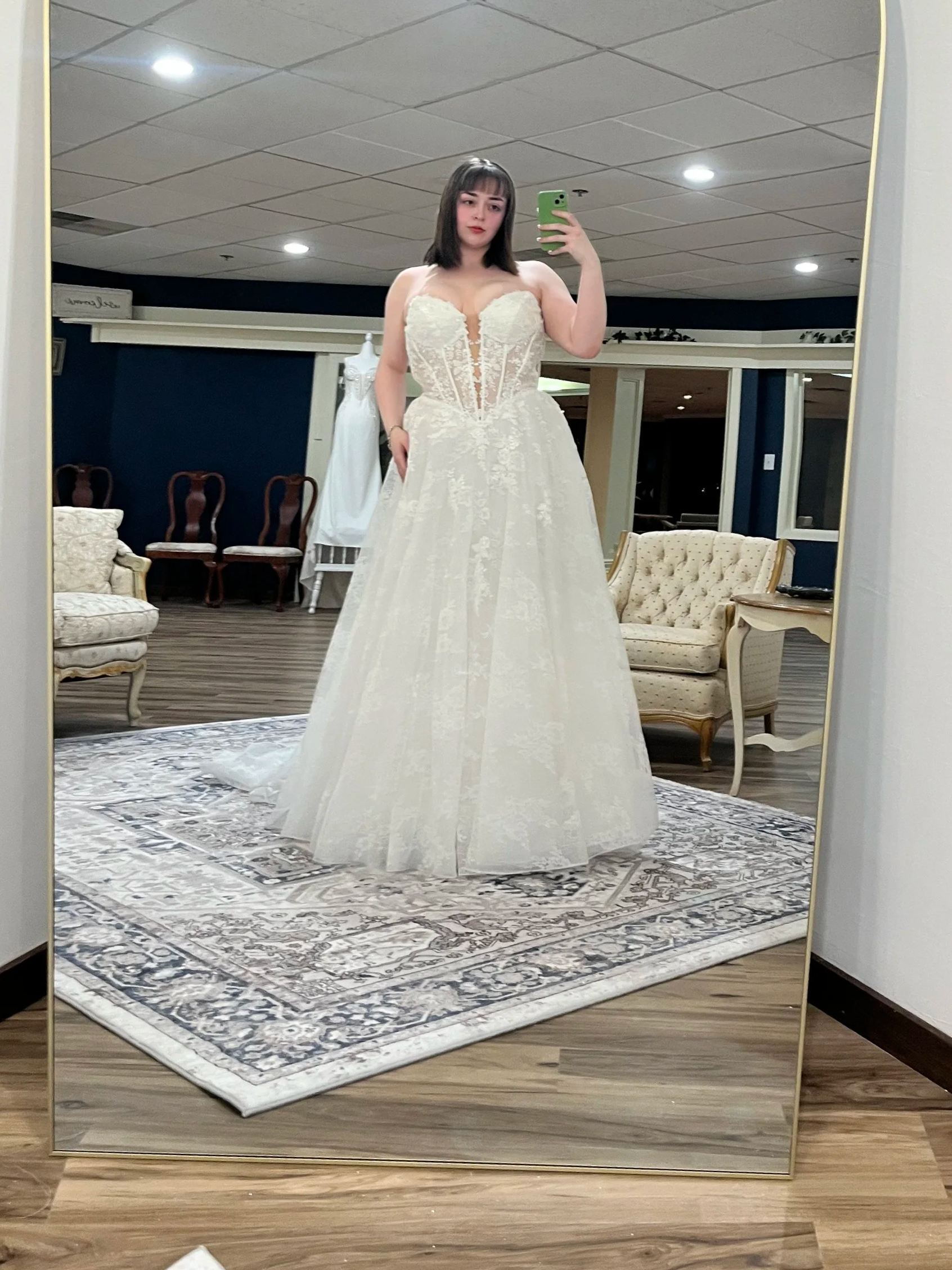 A woman trying on a white wedding dress in front of a mirror in a fitting room. The space is intentionally large and made to feel intimate for trying on wedding gowns.