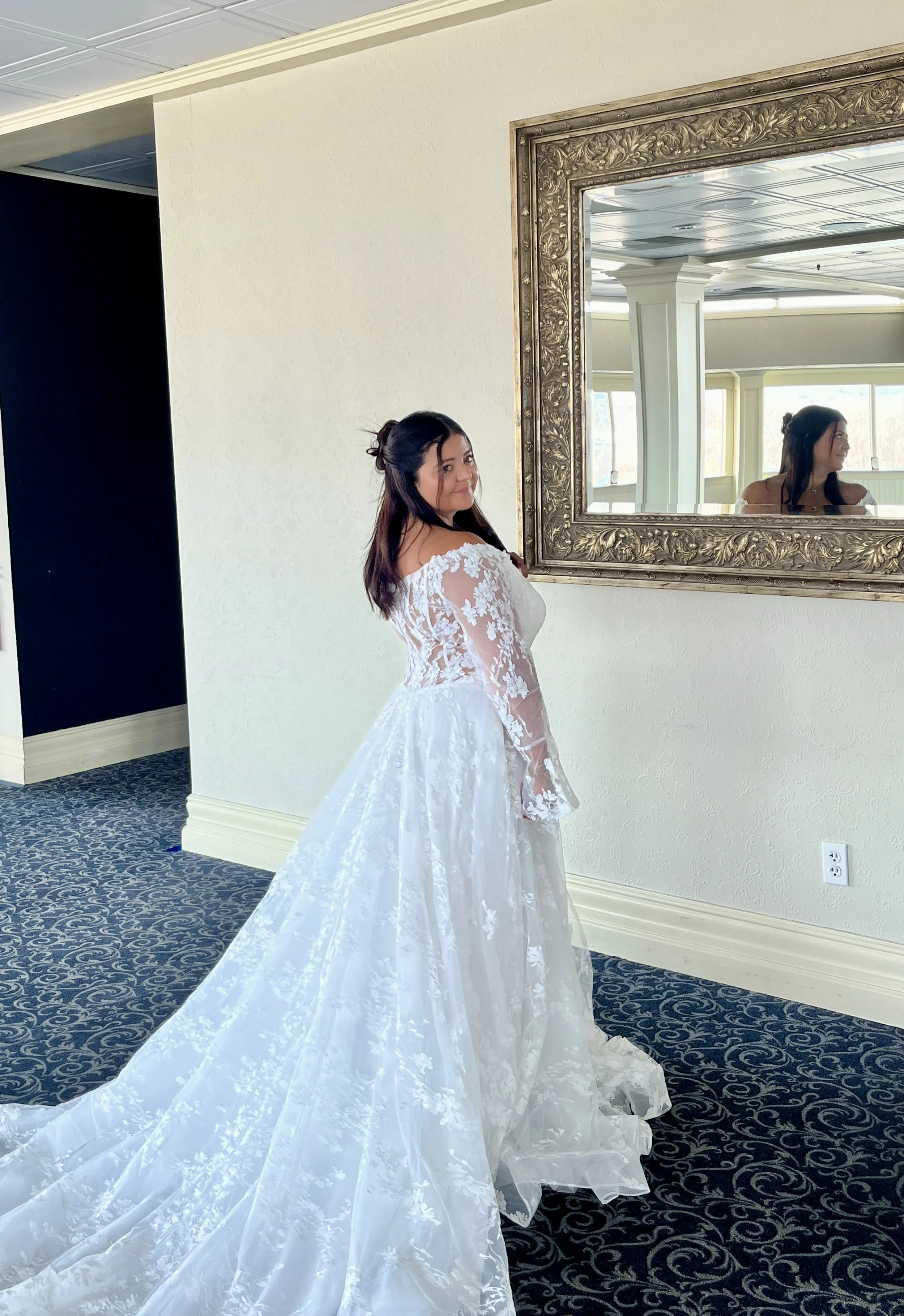 A woman in an ivory wedding gown with lace details stands in front of a large ornate mirror, smiling. Her long train is displayed beautifully.