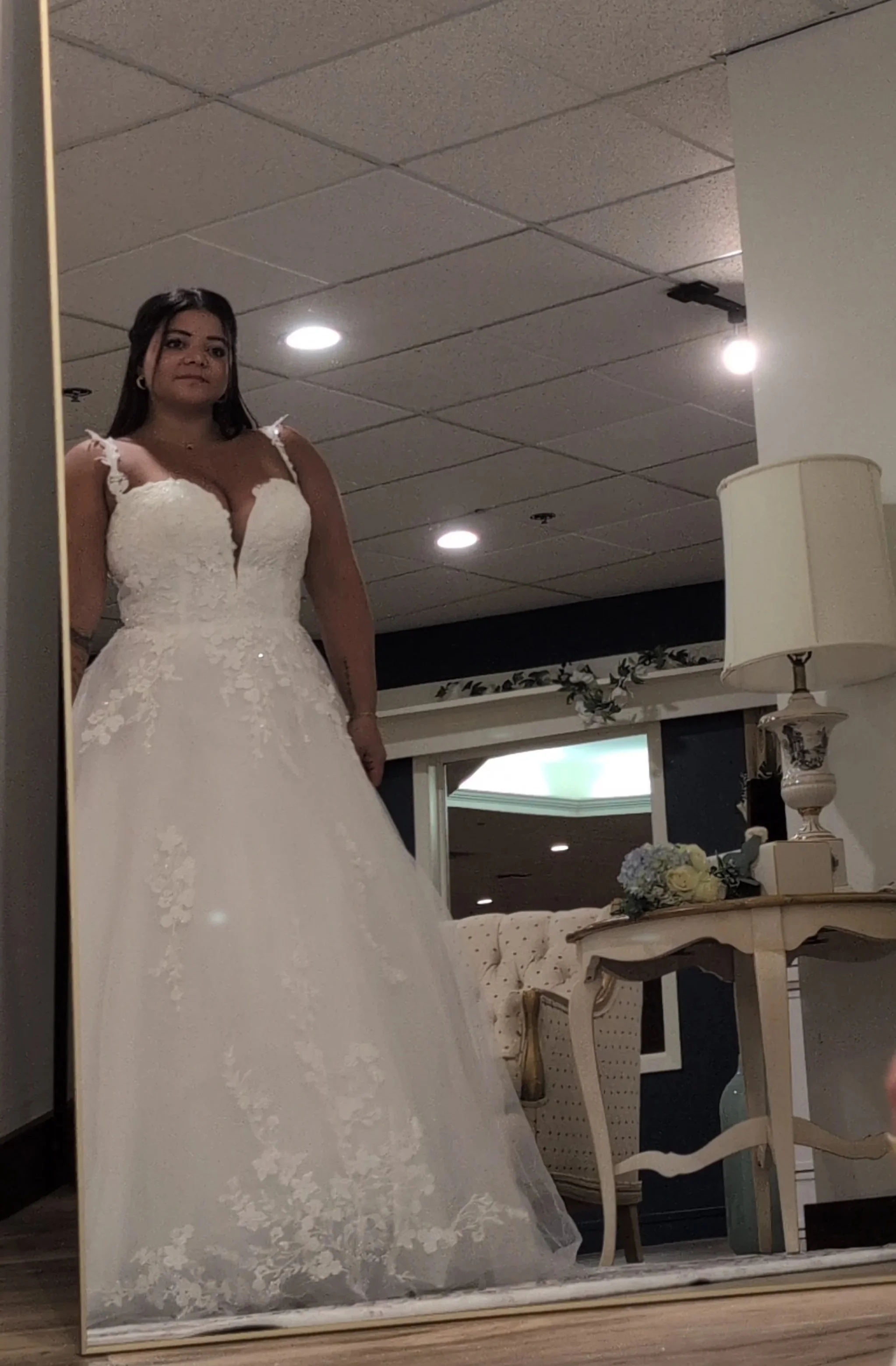 A woman taking a mirror selfie in a bridal boutique wearing a white wedding gown with lace details.