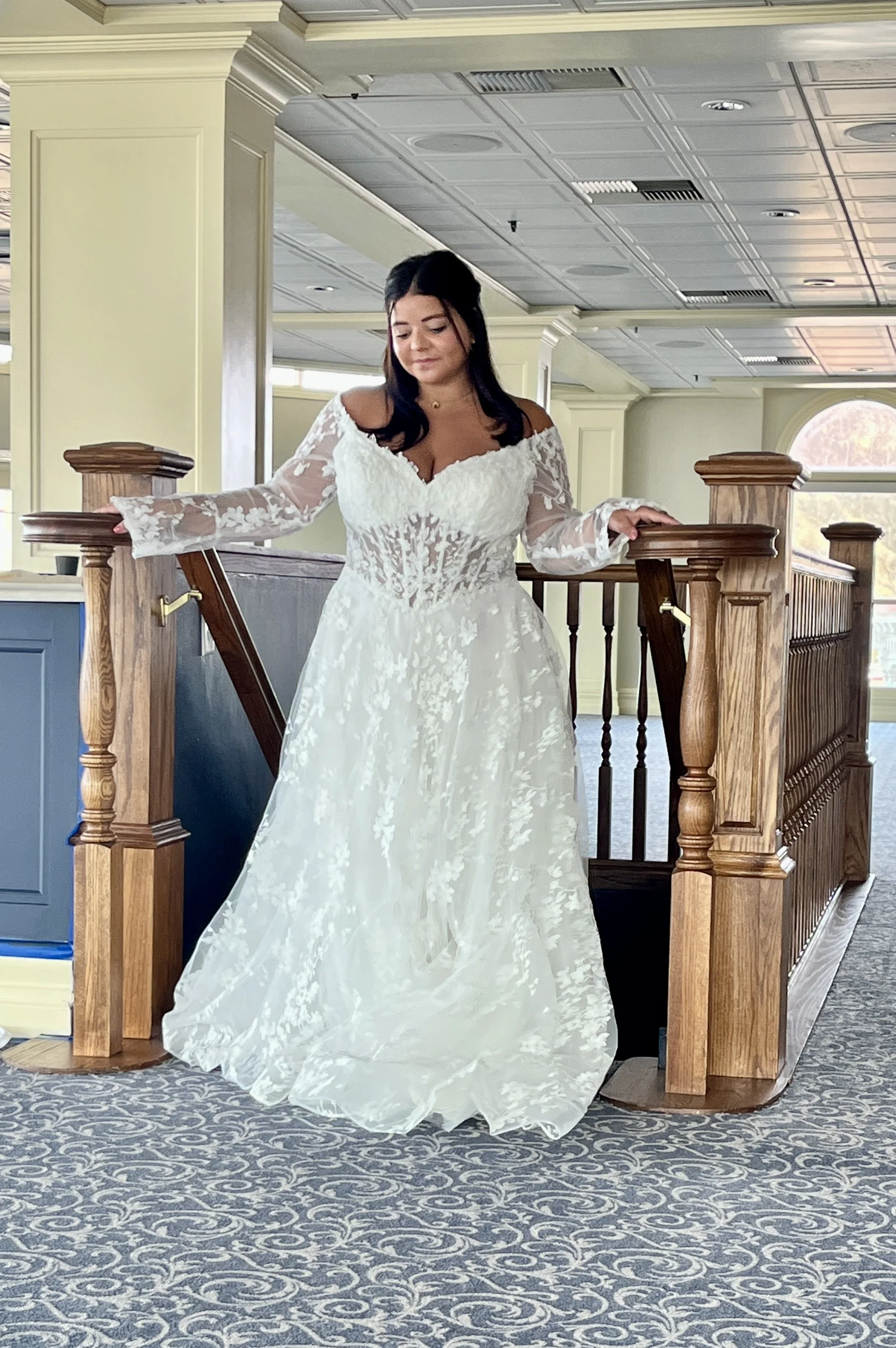 A woman in an ivory wedding dress standing on a staircase inside a building. The dress has long sleeves, a-line silhouette, and lots of floral lace appliques.