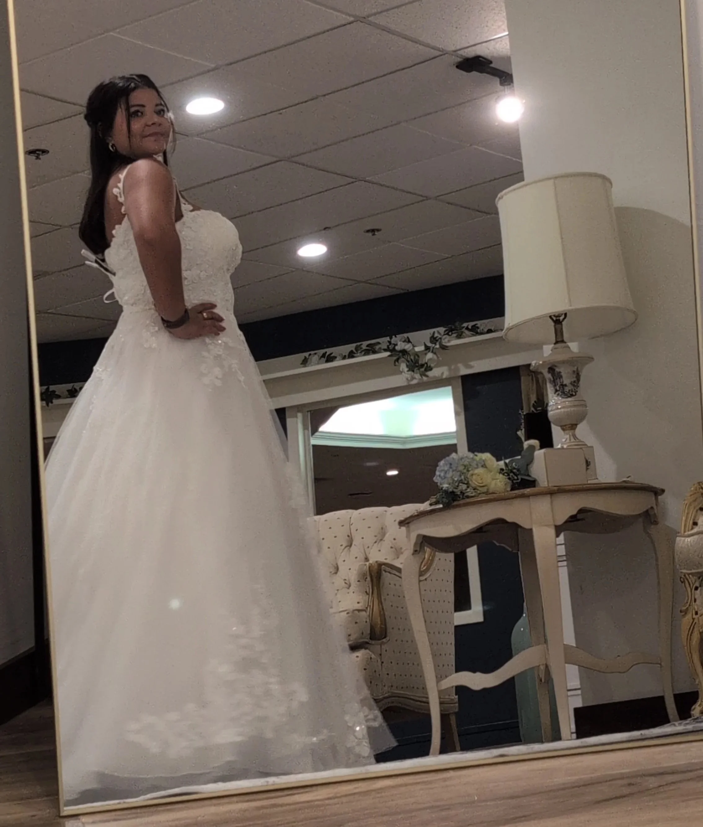 A woman in a white wedding dress taking a mirror selfie in a room with elegant furniture and a floral arrangement on a table.