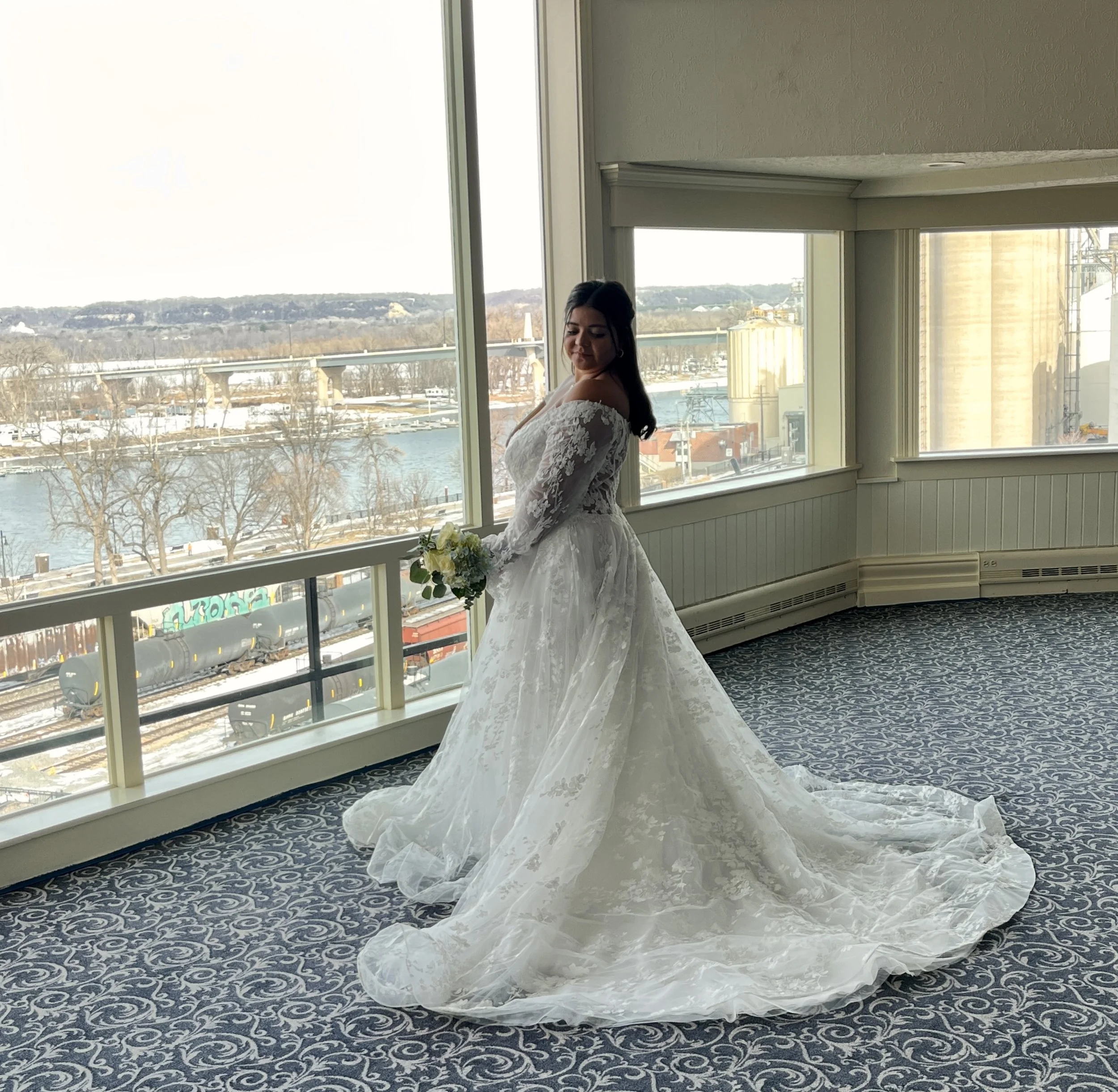 Brunette bride in a white lace wedding dress holding a bouquet of white and green flowers, standing near a large window with a river and industrial buildings outside. Ivory & Wing is located inside the St James Hotel, where this photo was taken.