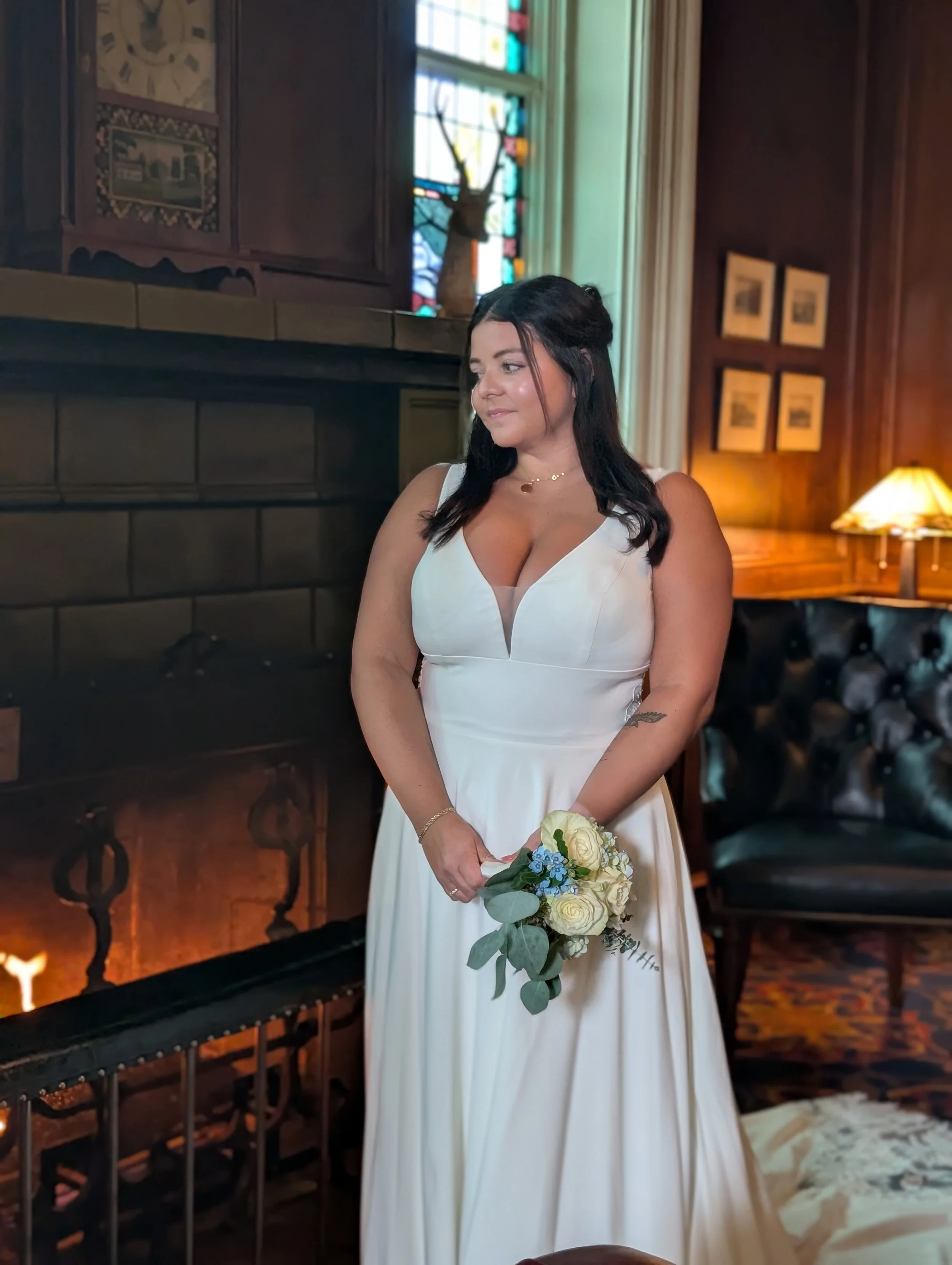 A woman in a white wedding dress holding a bouquet indoors with a stained glass window and wooden paneling in the background.
