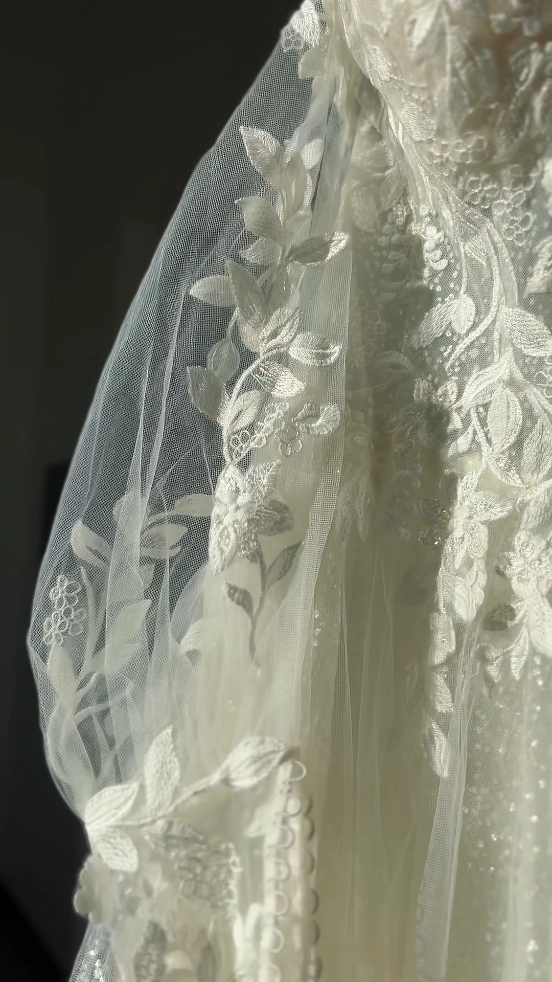 Close-up of a wedding dress' detachable sleeve with intricate floral embroidery and lace detail, illuminated by sunlight.