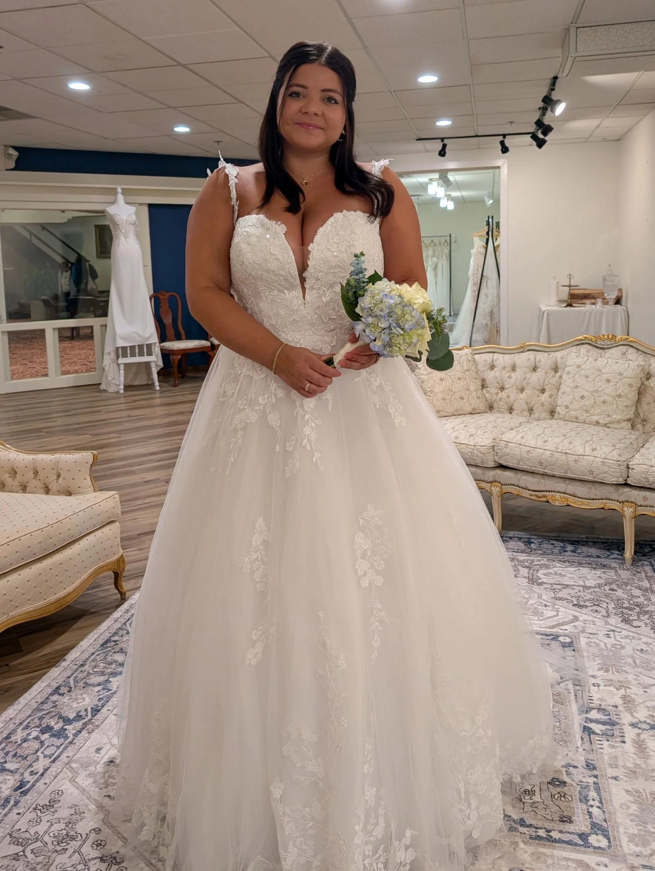 A woman in a wedding dress holding a bouquet of white and blue flowers in Ivory & Wing Bridal boutique with vintage furniture and wedding dresses in the background. The flowers are from Clementine, another business located inside the St James Hotel.